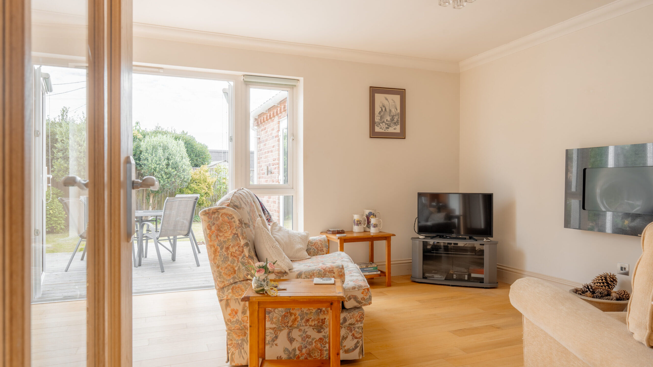Bright living room with wooden floor, floral armchair, beige sofa, small tables, flat-screen TV on a stand, and open glass doors leading to a patio with outdoor furniture and a garden view.