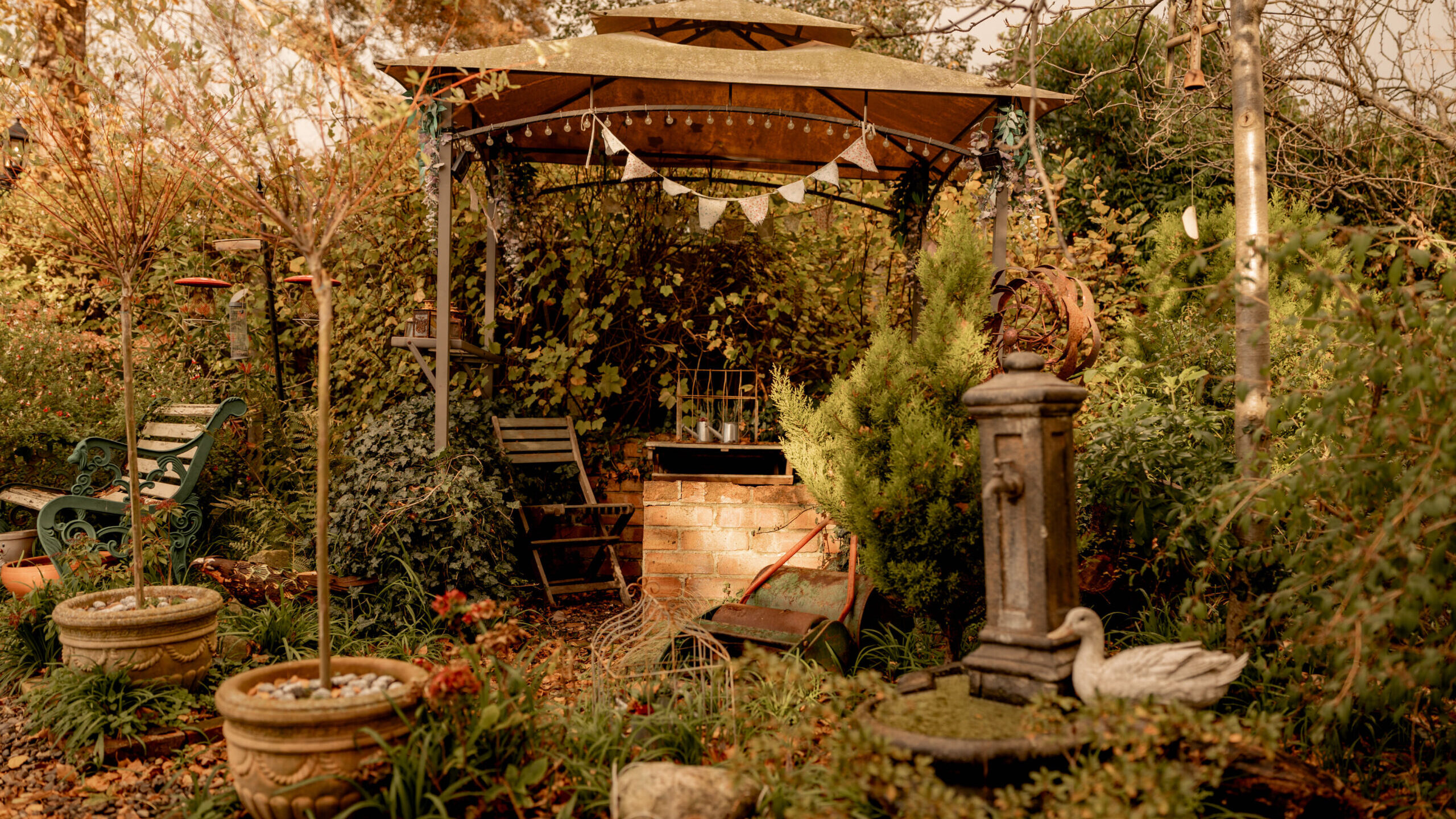 A cozy garden scene with a wooden gazebo surrounded by lush greenery, potted plants, and fallen leaves. There are chairs under the gazebo and a decorative fountain with a bird statue in the foreground.