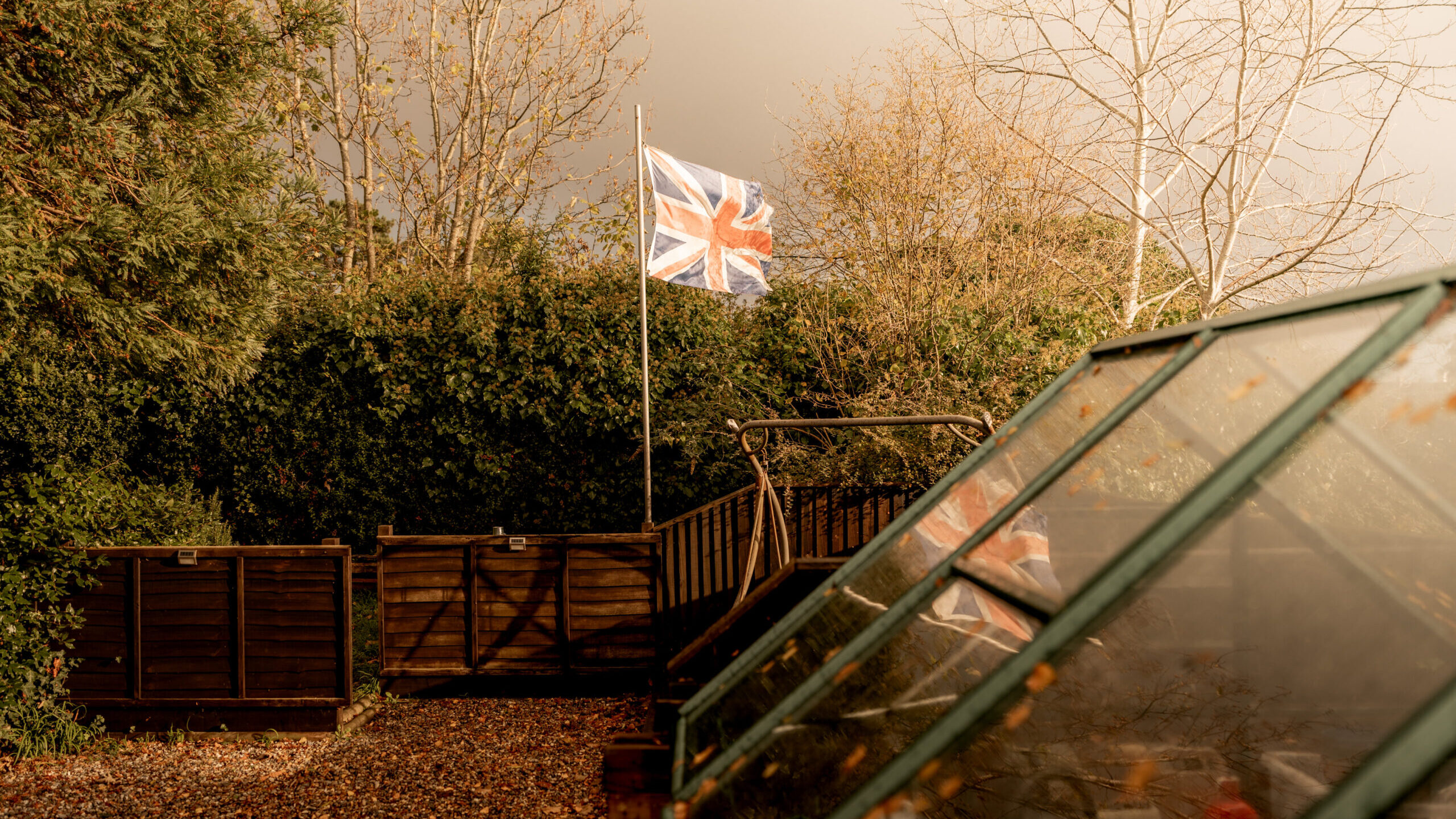 A Union Jack flag flies on a pole in a garden surrounded by autumn trees, with a greenhouse in the foreground and wooden compost bins in the background.