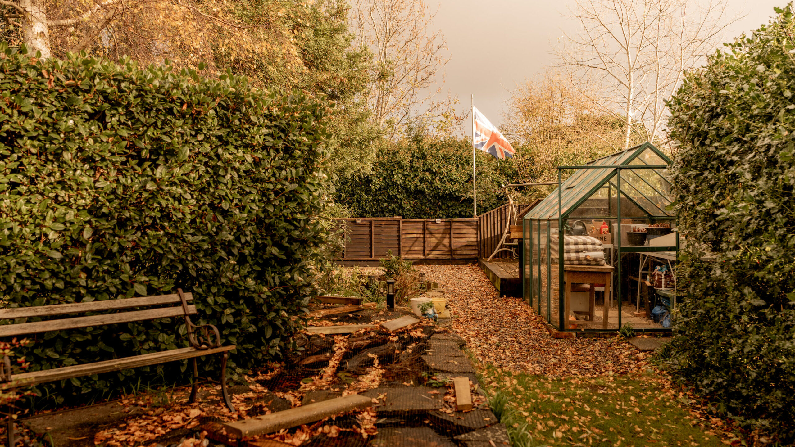 A garden scene with a bench, overgrown plants, fallen leaves, a wooden path, and a glass greenhouse. In the background, a flag with the Union Jack is raised near a fence under autumn trees.