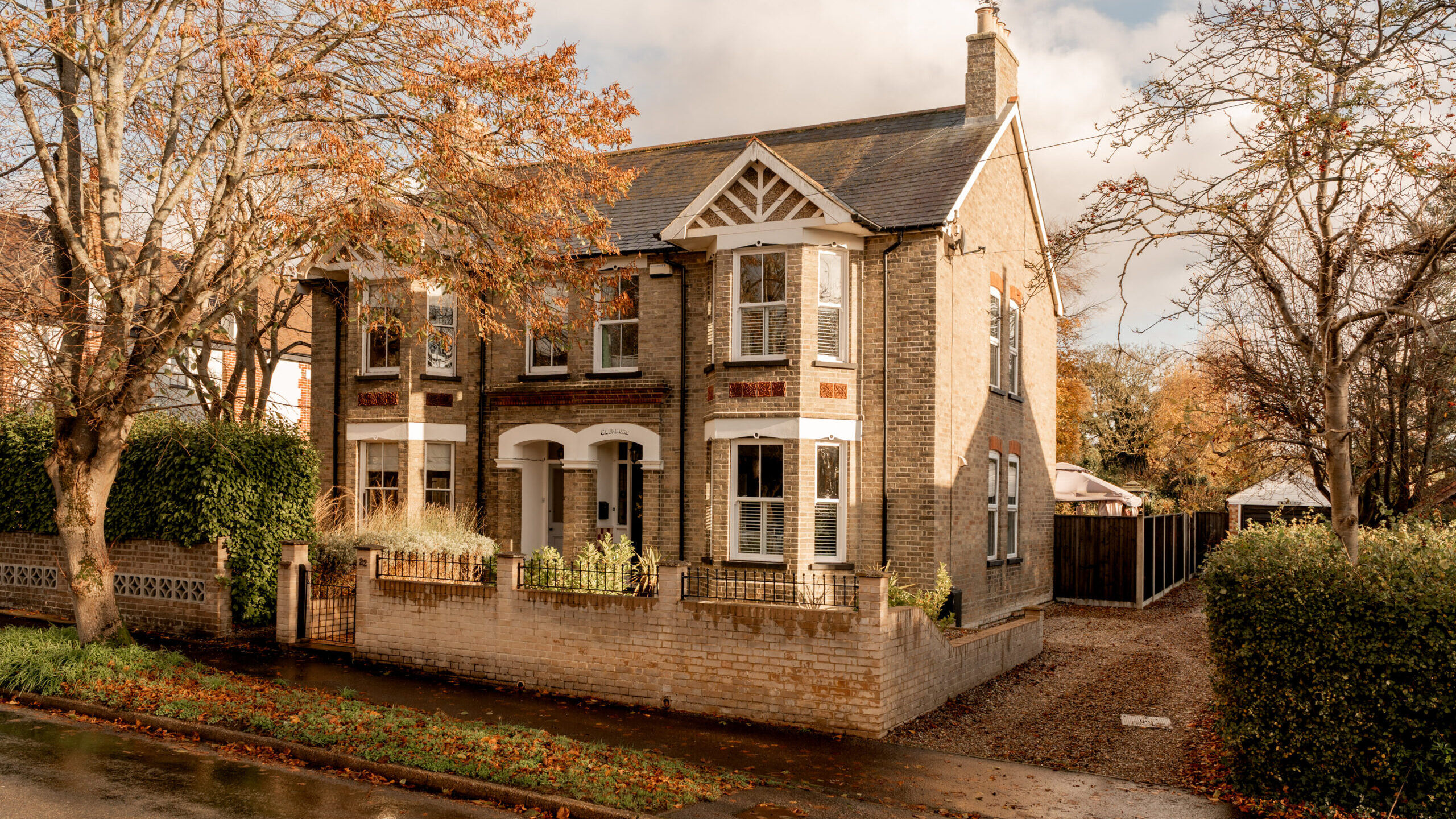A two-story brick house with white trim, bay windows, and a small front garden, surrounded by autumn trees. A driveway runs beside the house, and wet pavement lines the street in front.