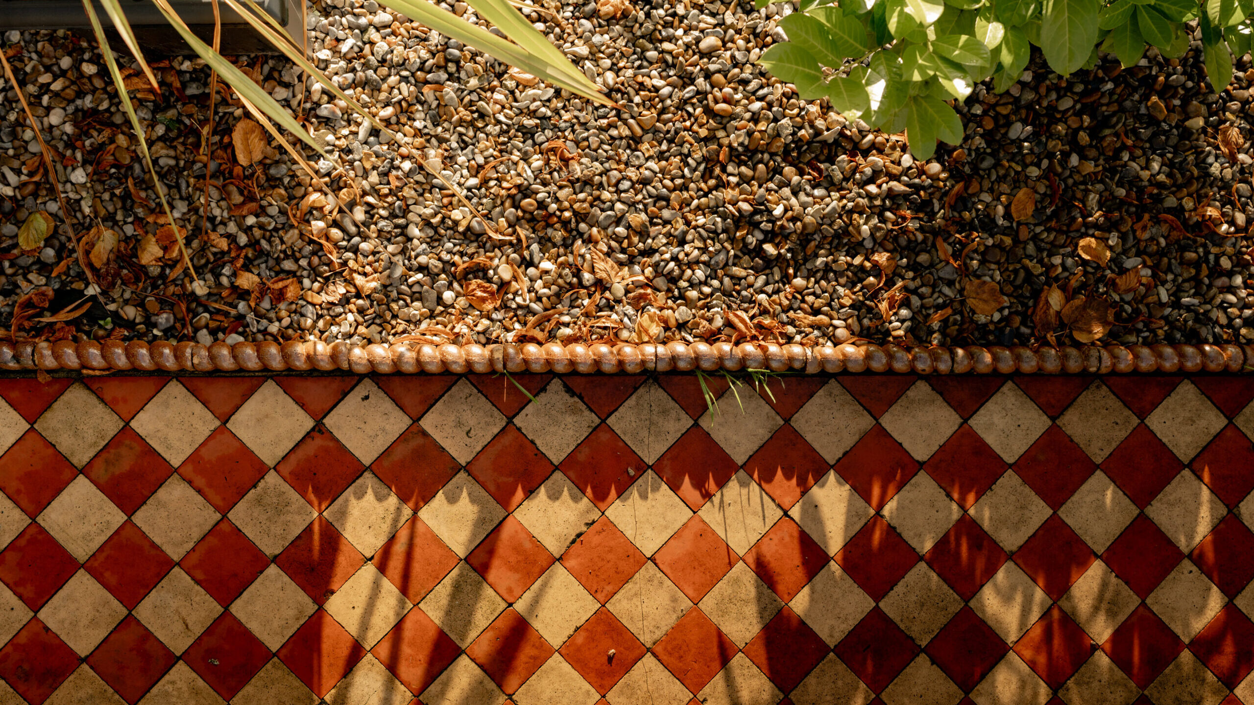 Top view of a garden bed with pebbles and green plants above a red and beige diamond-patterned tile floor. Sunlight casts plant shadows onto the tiles.