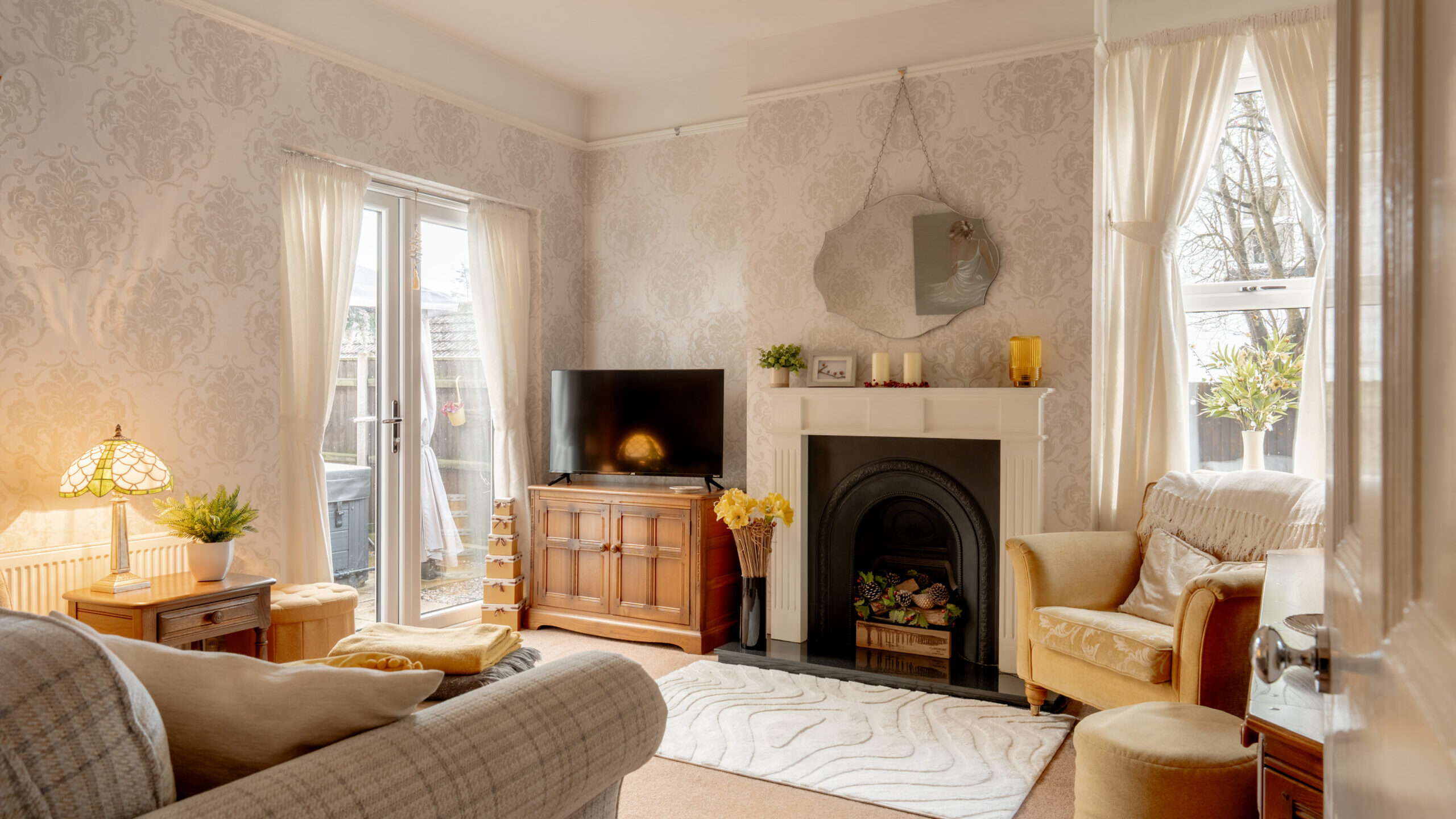 A cozy living room with patterned wallpaper, a fireplace, a TV on a wooden stand, a beige sofa, two armchairs, and large windows with sheer curtains letting in natural light.