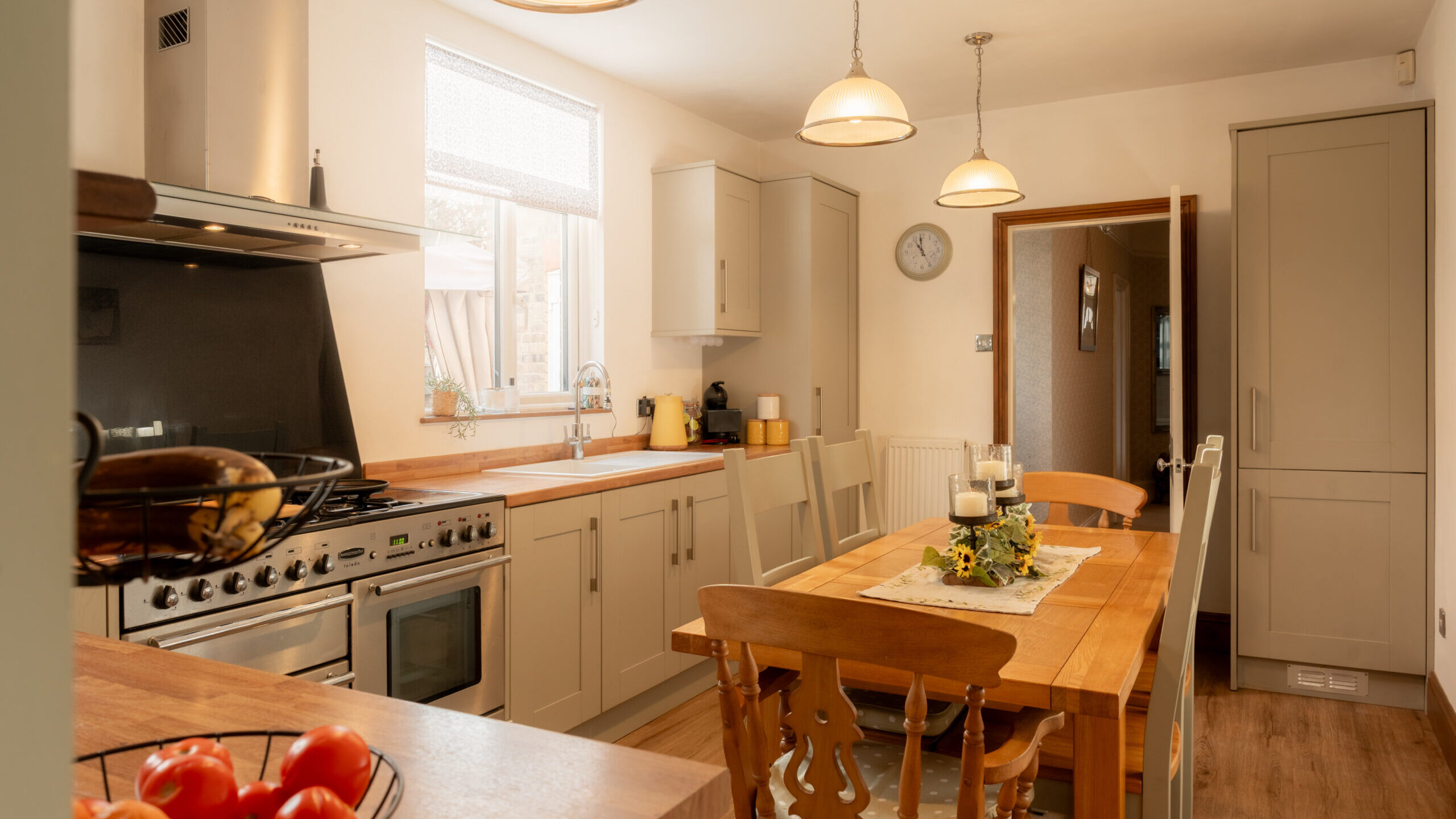 A cozy, well-lit kitchen with beige cabinets, a wooden dining table with four chairs, a modern gas stove, fruit bowl, and pendant lights hanging from the ceiling. A window brings in natural light.