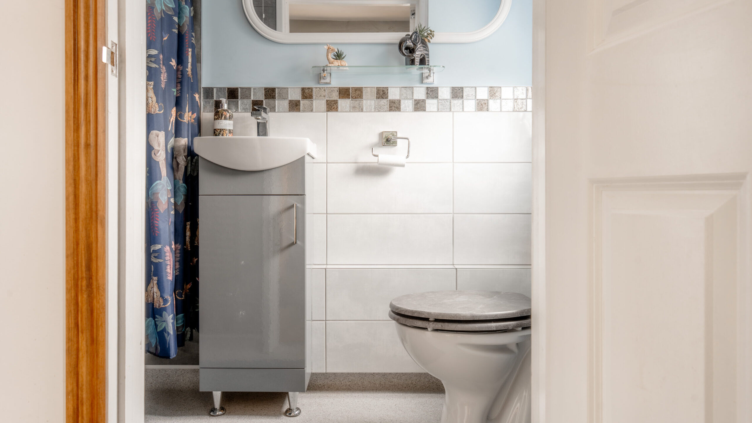 A small bathroom with a white toilet, a gray vanity with a sink, a rectangular mirror with a white frame, a shelf with small plants, and a blue shower curtain with animal prints. The walls are blue and white tiled.