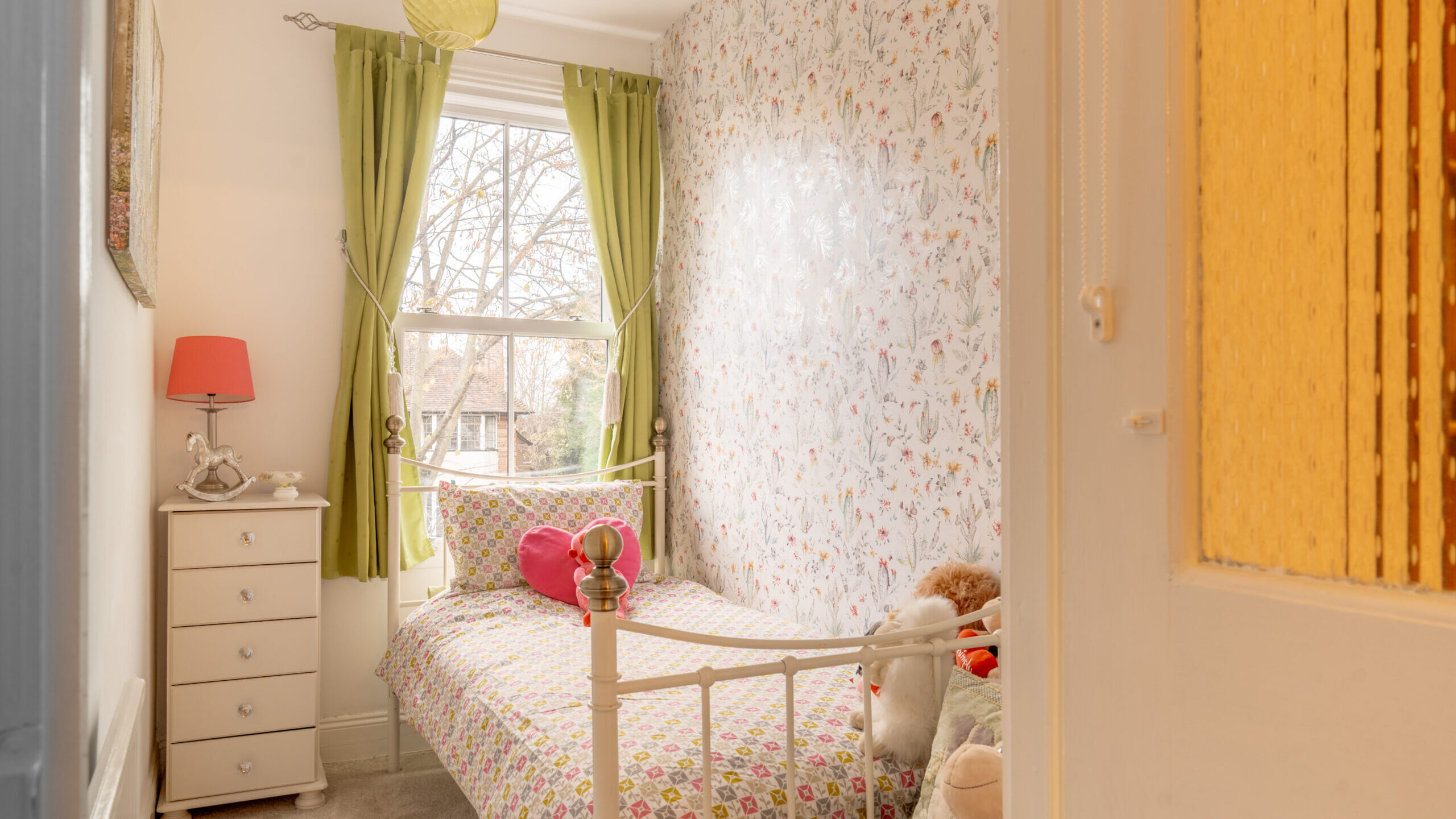 A cozy, small bedroom with floral wallpaper, a single bed with colorful bedding and a heart-shaped pillow, green curtains, a white dresser with a red lamp, and stuffed animals on the bed. Sunlight shines through the window.