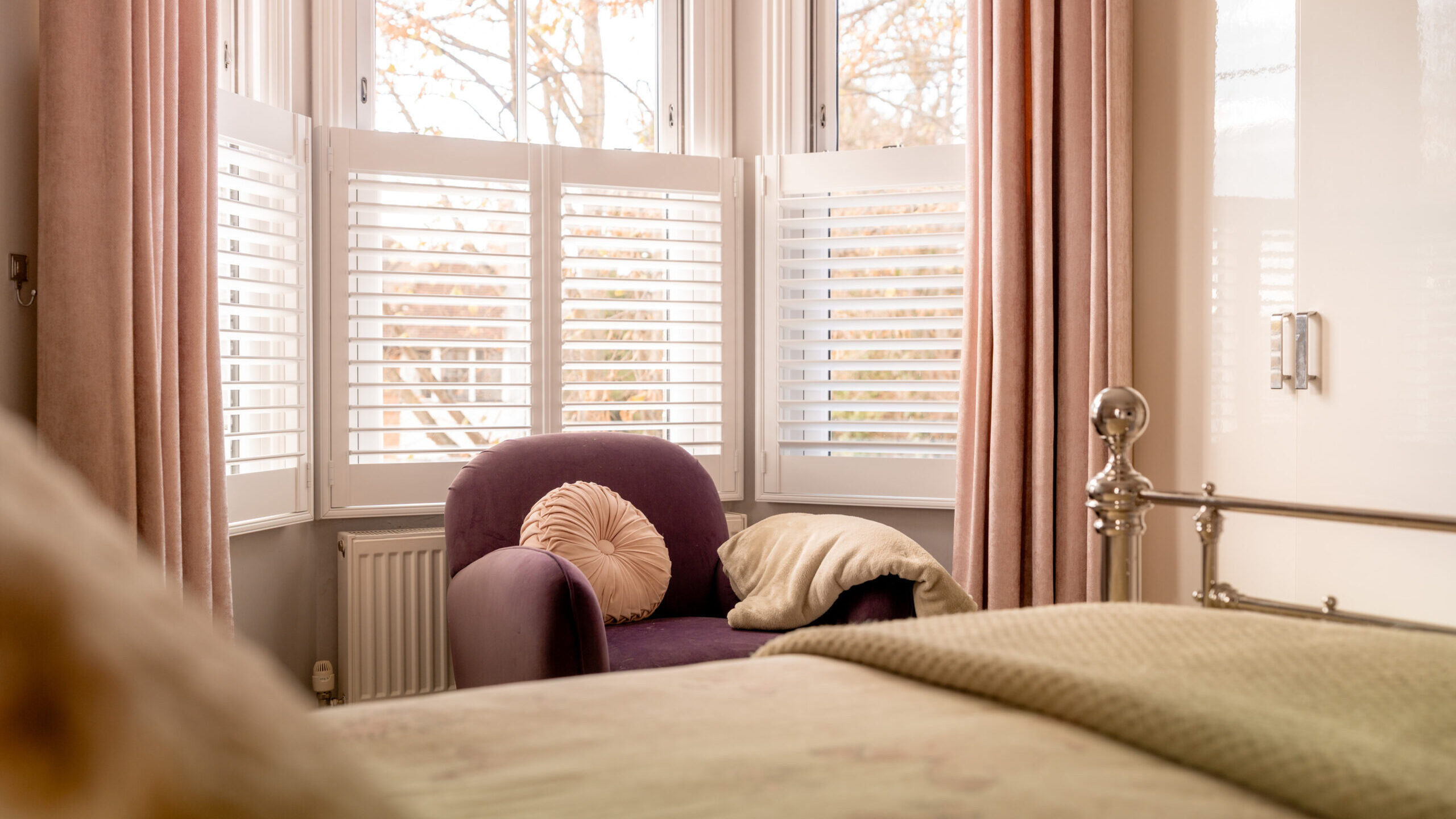 A cozy bedroom with a purple armchair by a bay window with white shutters, pink curtains, a round cushion, and a blanket. Part of a bed with a greenish cover is visible in the foreground.