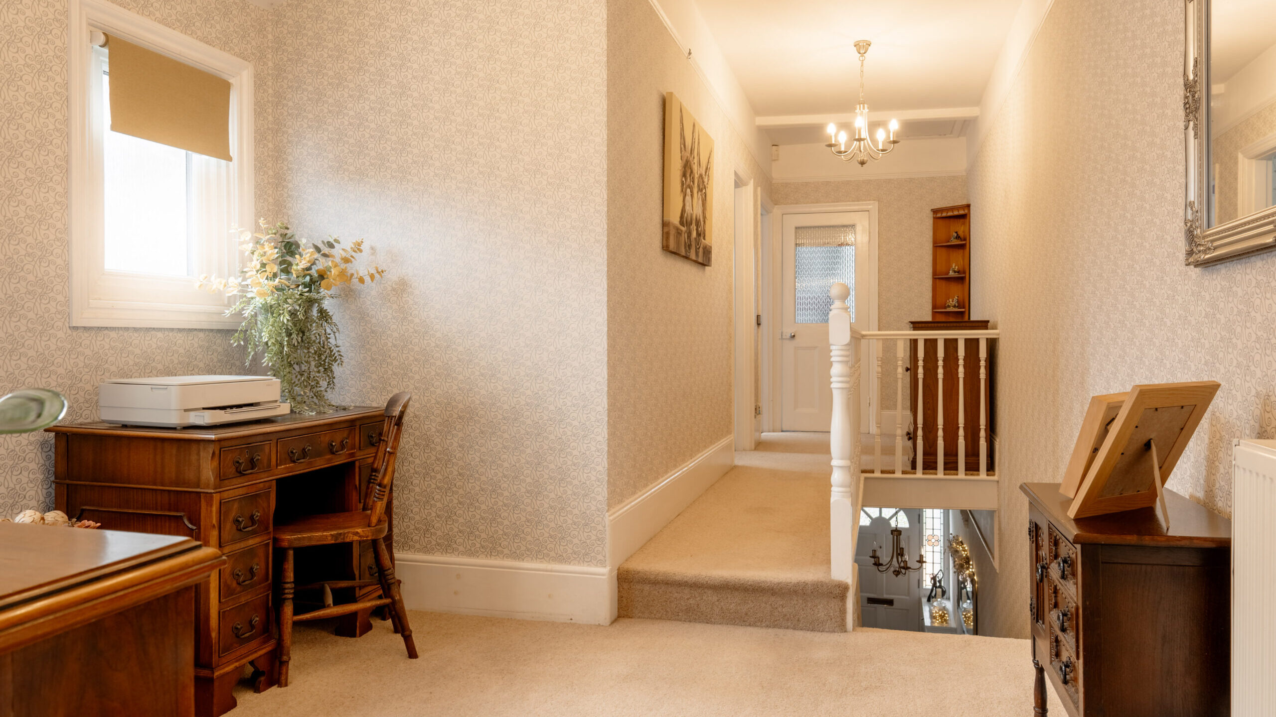 A bright hallway with beige carpet, patterned wallpaper, and wooden furniture, including a desk with a printer, a chest of drawers, and a mirror. A staircase with white railing leads downstairs. Flowers sit on the desk.