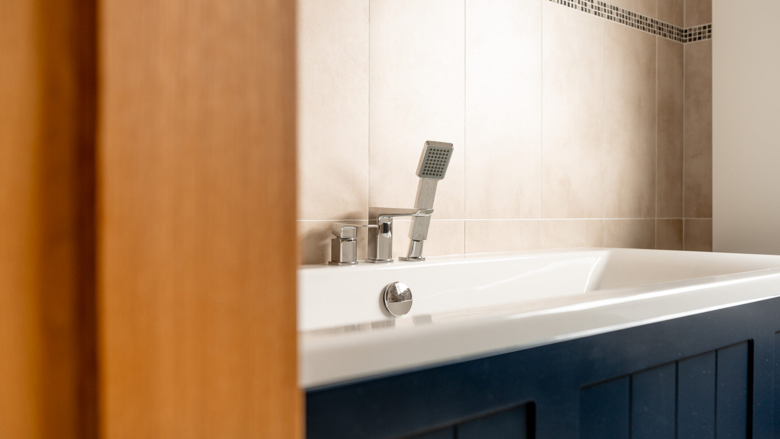 Close-up of a modern bathtub with a blue wooden panel, chrome fixtures, and a handheld showerhead, set against beige tiled walls with a decorative mosaic tile strip near the top.