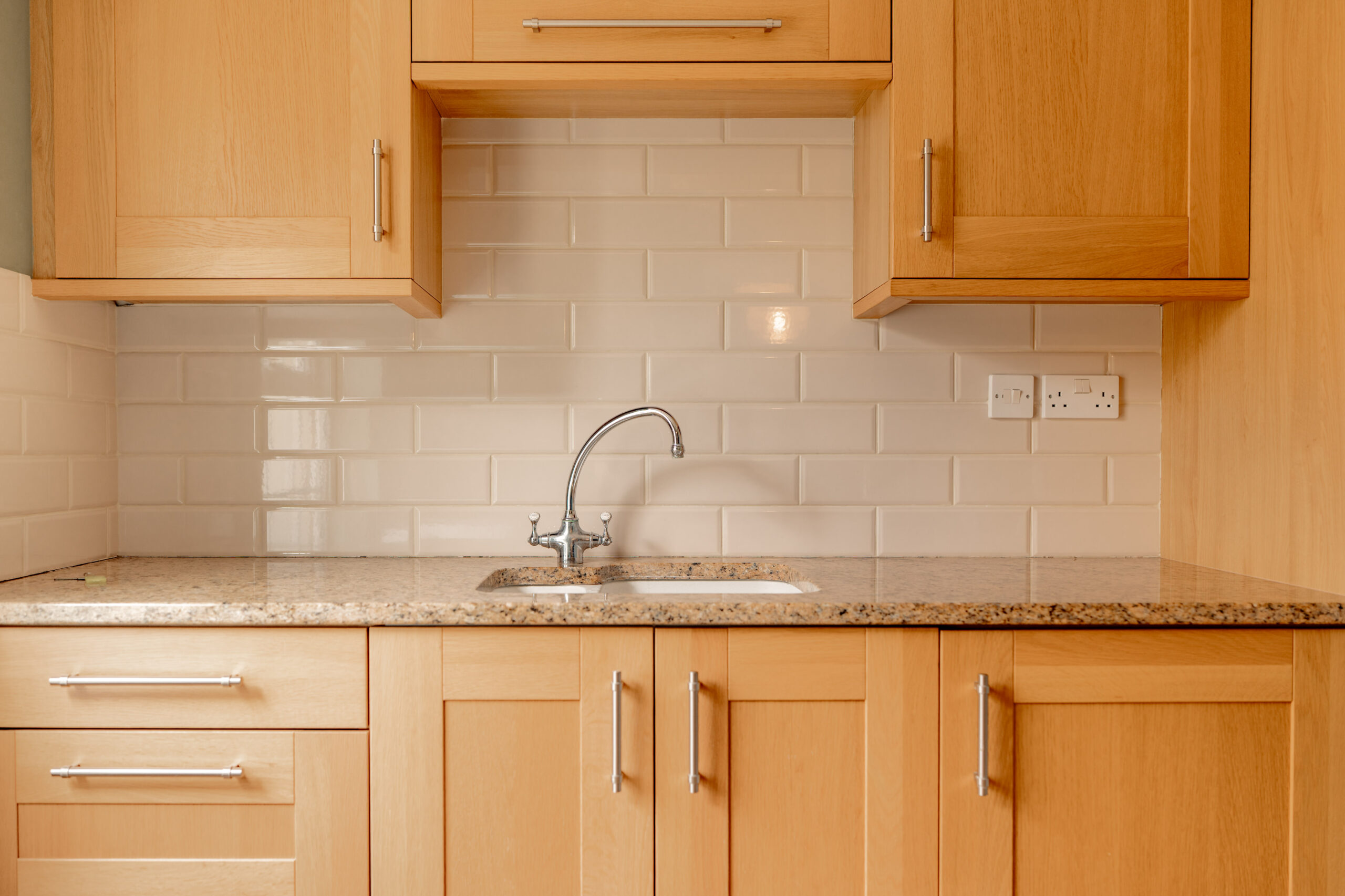 A small kitchen area with light wood cabinets, a granite countertop, a stainless steel faucet over a small sink, and a white subway tile backsplash with two electrical outlets.