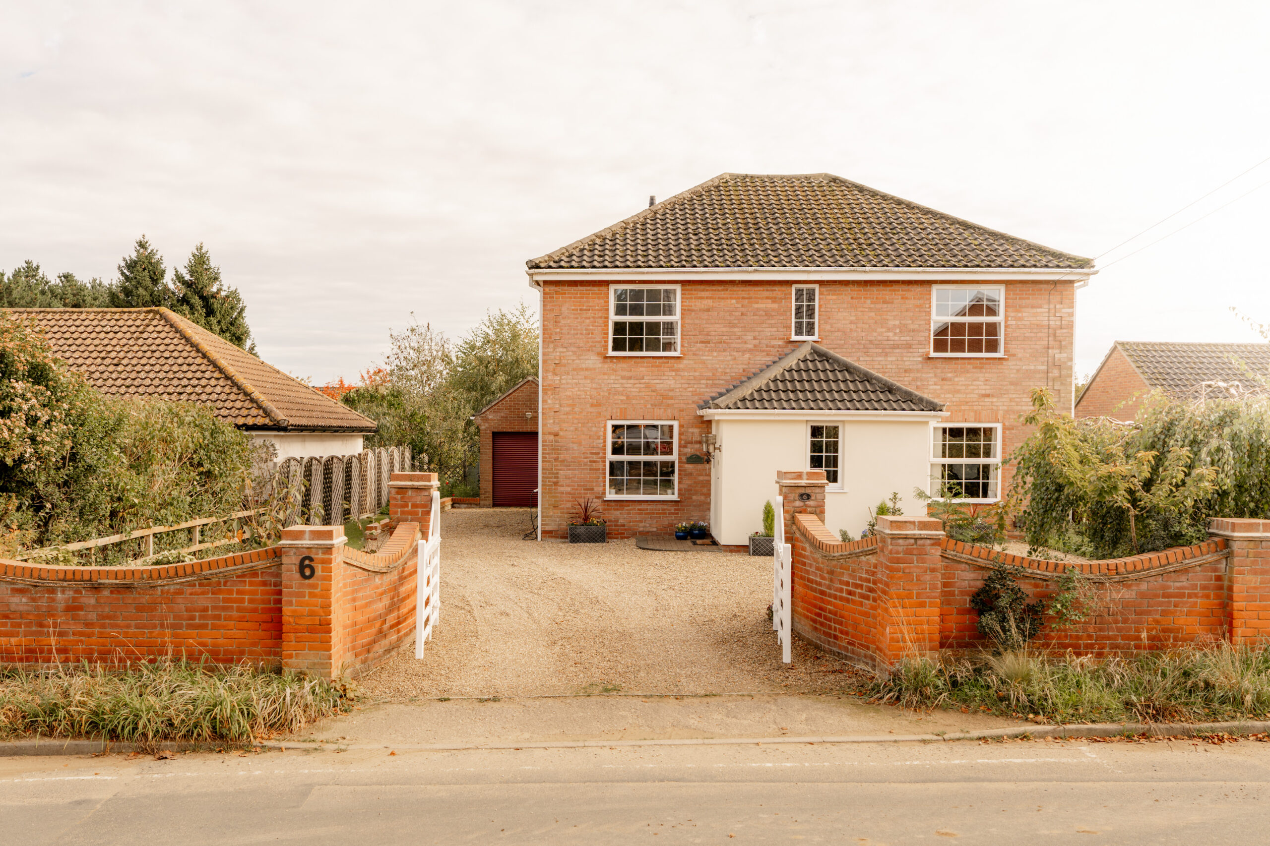 A two-story brick house with a tiled roof, white-framed windows, and a gravel driveway, surrounded by a low red brick wall and white gates, with greenery and trees nearby. The house number 6 is on the wall.