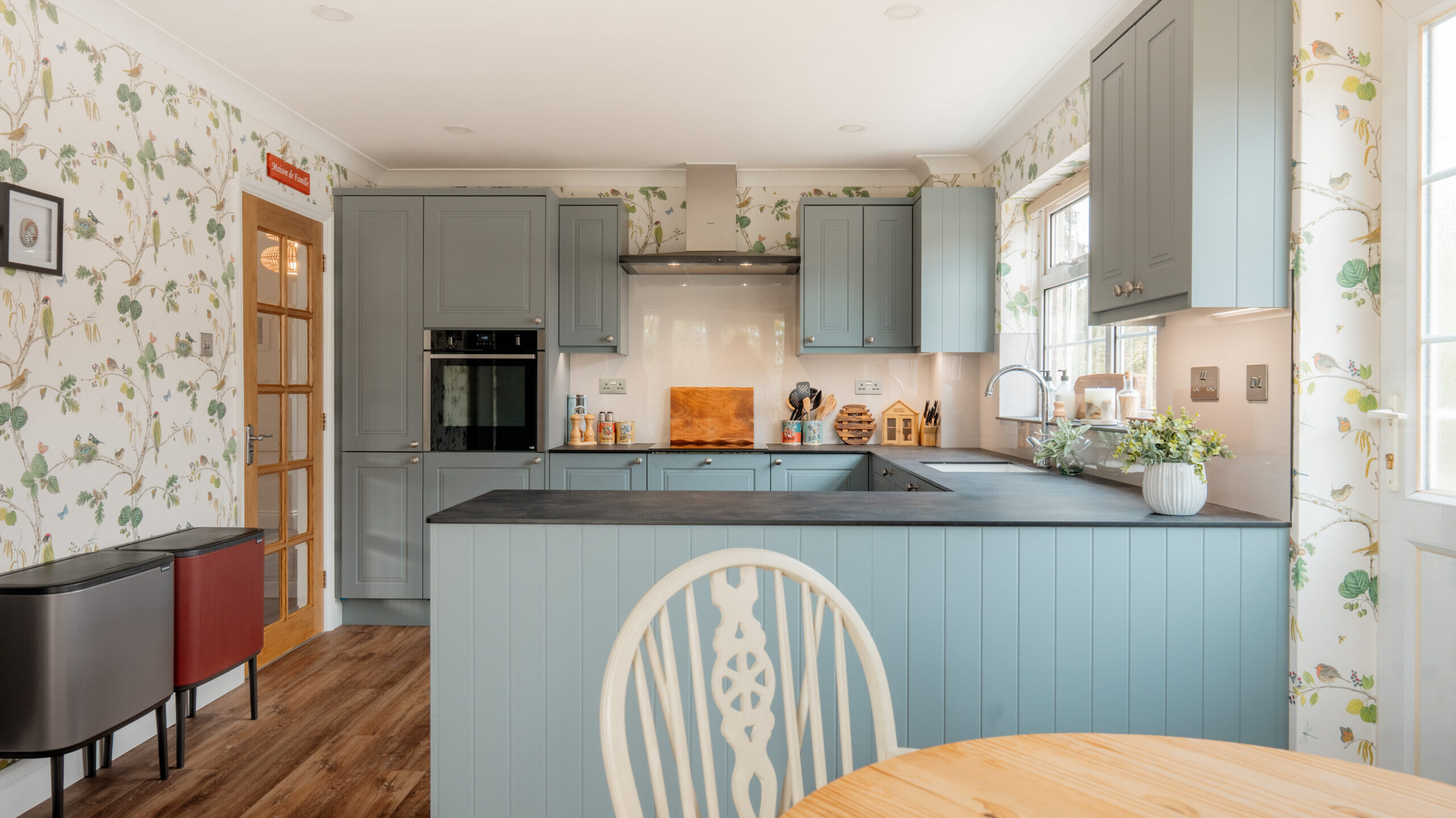 A cozy kitchen with light blue cabinets, a black countertop, floral wallpaper, wooden floor, and a round wooden table with a decorative white chair in the foreground. Sunlight streams through the window.