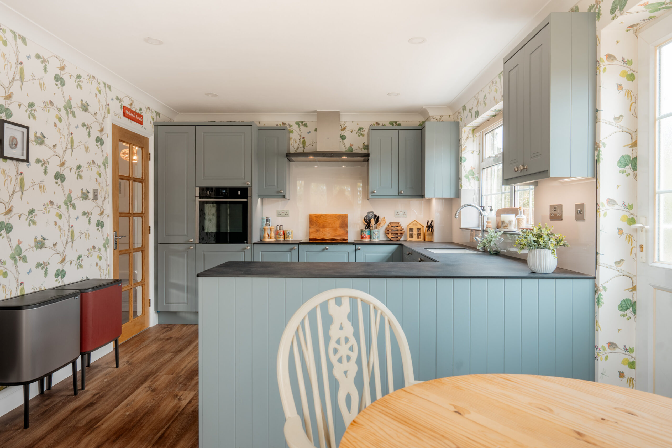 A cozy kitchen with light blue cabinets, a black countertop, floral wallpaper, wooden floor, and a round wooden table with a decorative white chair in the foreground. Sunlight streams through the window.
