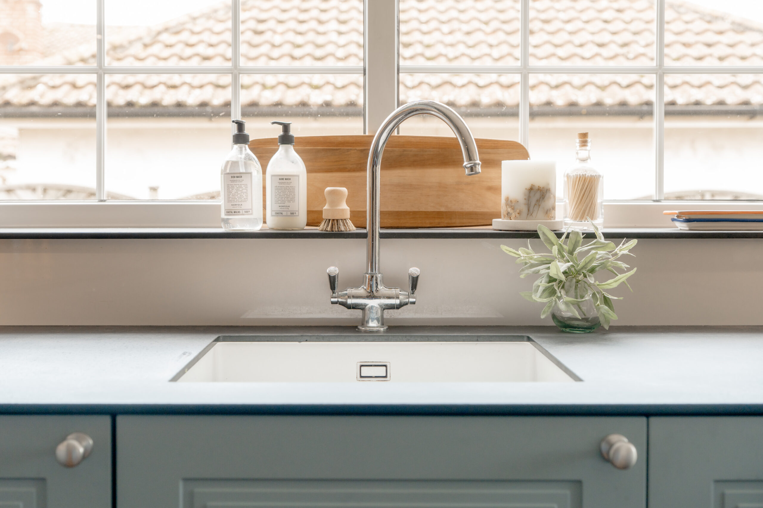 A modern kitchen sink with a silver faucet, soap dispensers, a wooden dish brush, and a potted plant on the countertop, set in front of a large window with a tiled roof visible outside.