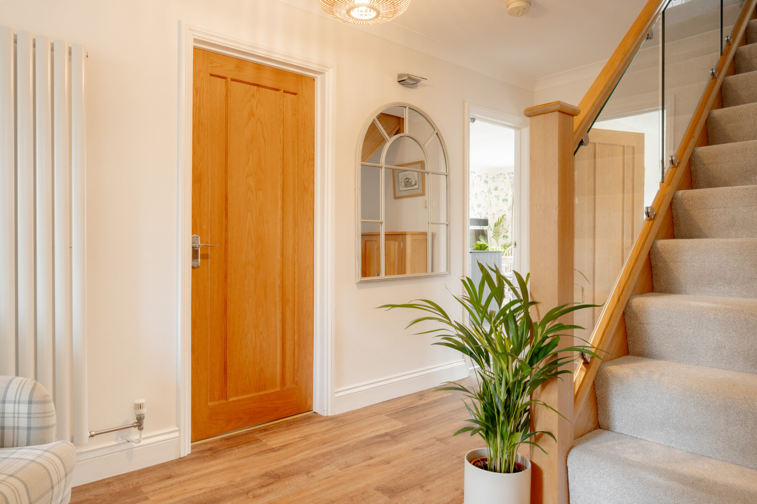 Bright hallway with wooden floor, closed wooden door, large decorative mirror, potted plant, staircase with glass balustrade, and a glimpse into an adjoining room with floral wallpaper.