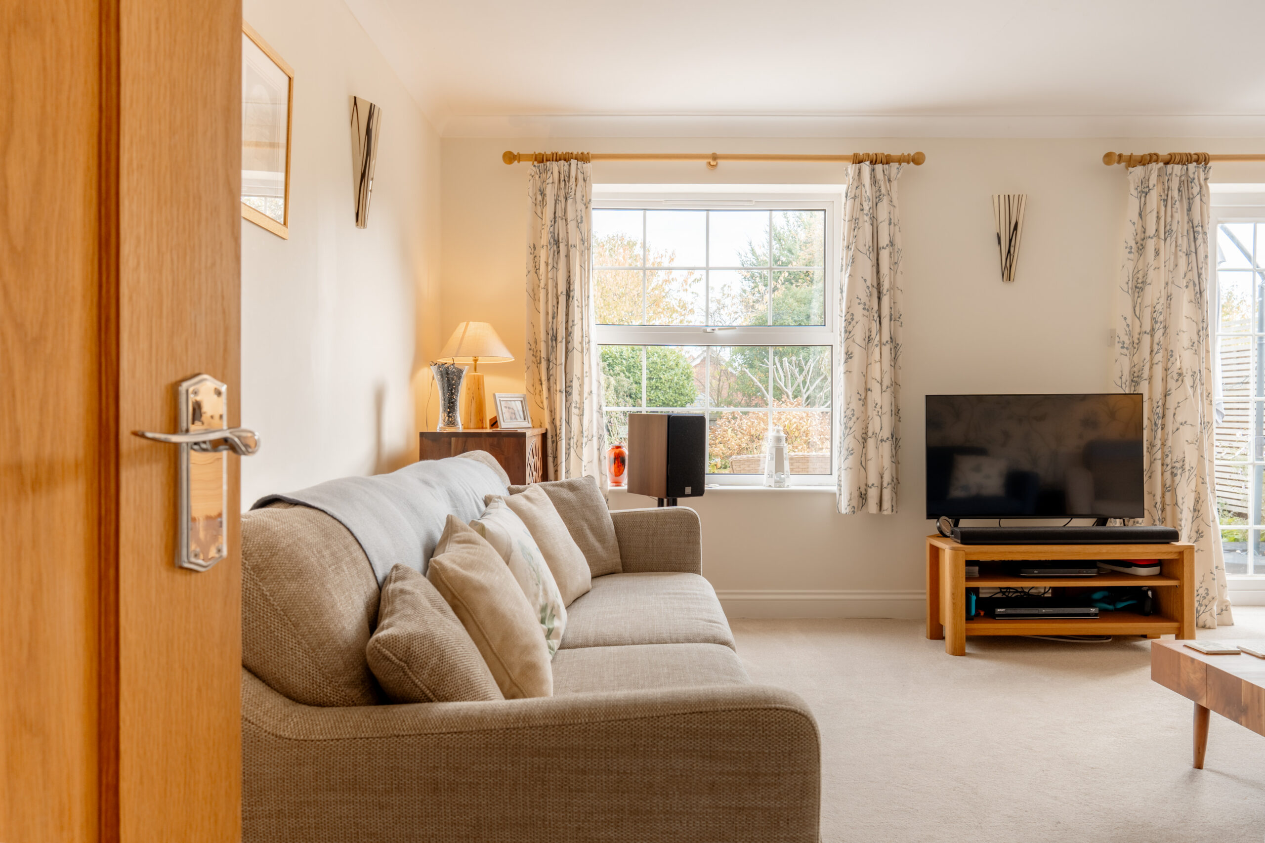 A cozy living room with a beige sofa, wooden furniture, a TV on a stand, and large windows with patterned curtains letting in natural light. There are decorative items and greenery visible outside.