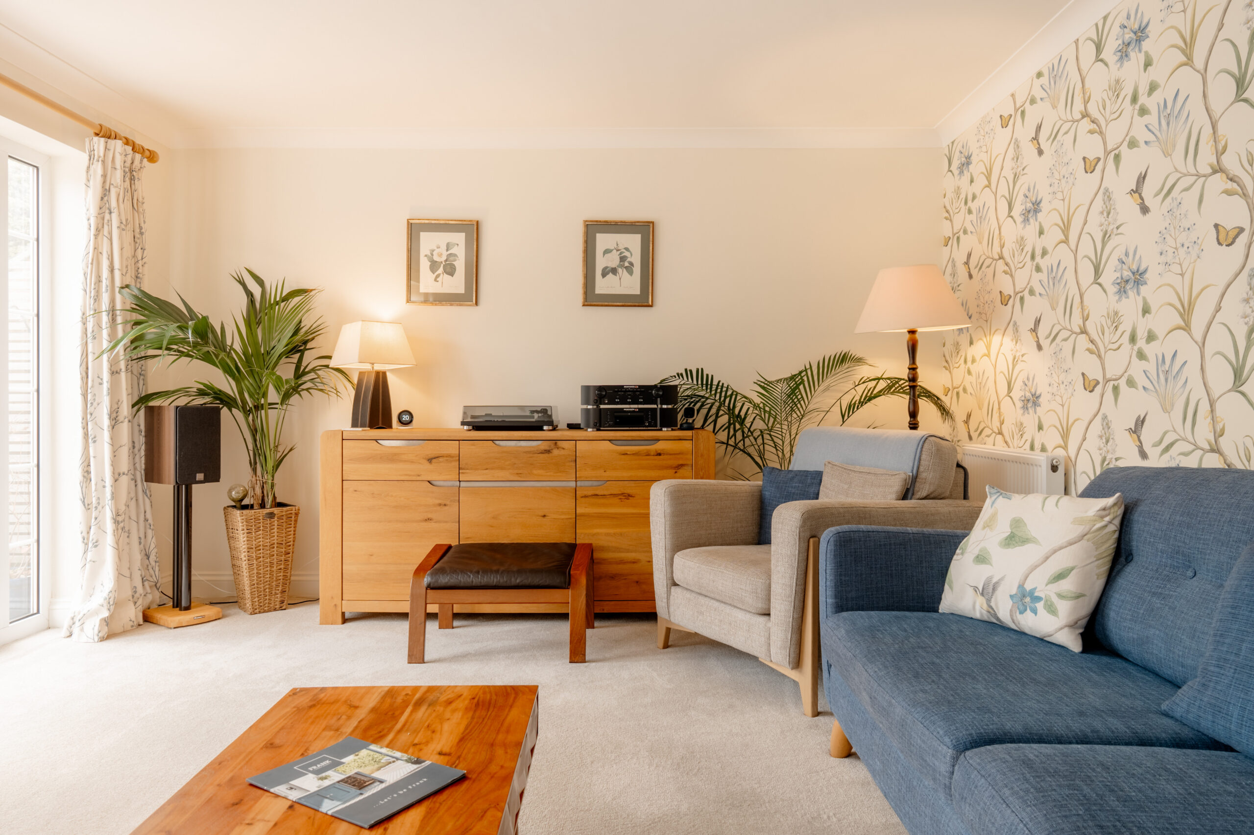 A cozy living room with a blue sofa, beige armchair, wooden sideboard, and coffee table. There are potted plants, lamps, framed wall art, and floral wallpaper, with sunlight coming through glass doors on the left.