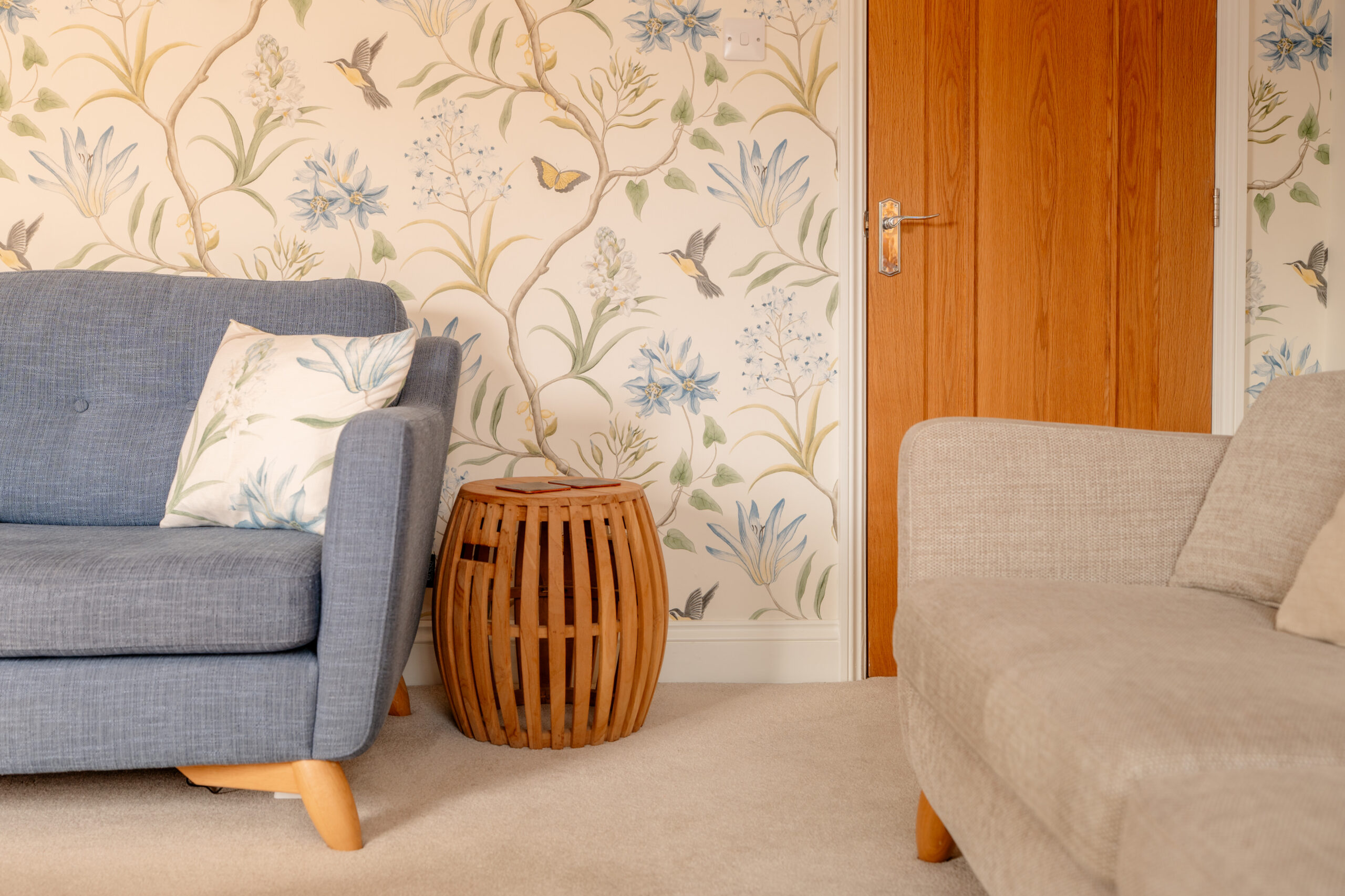 A cozy living room with a blue armchair, a beige sofa, a wooden side table, and wallpaper featuring floral and bird patterns. A wooden door is closed in the background. A white pillow with botanical prints is on the chair.