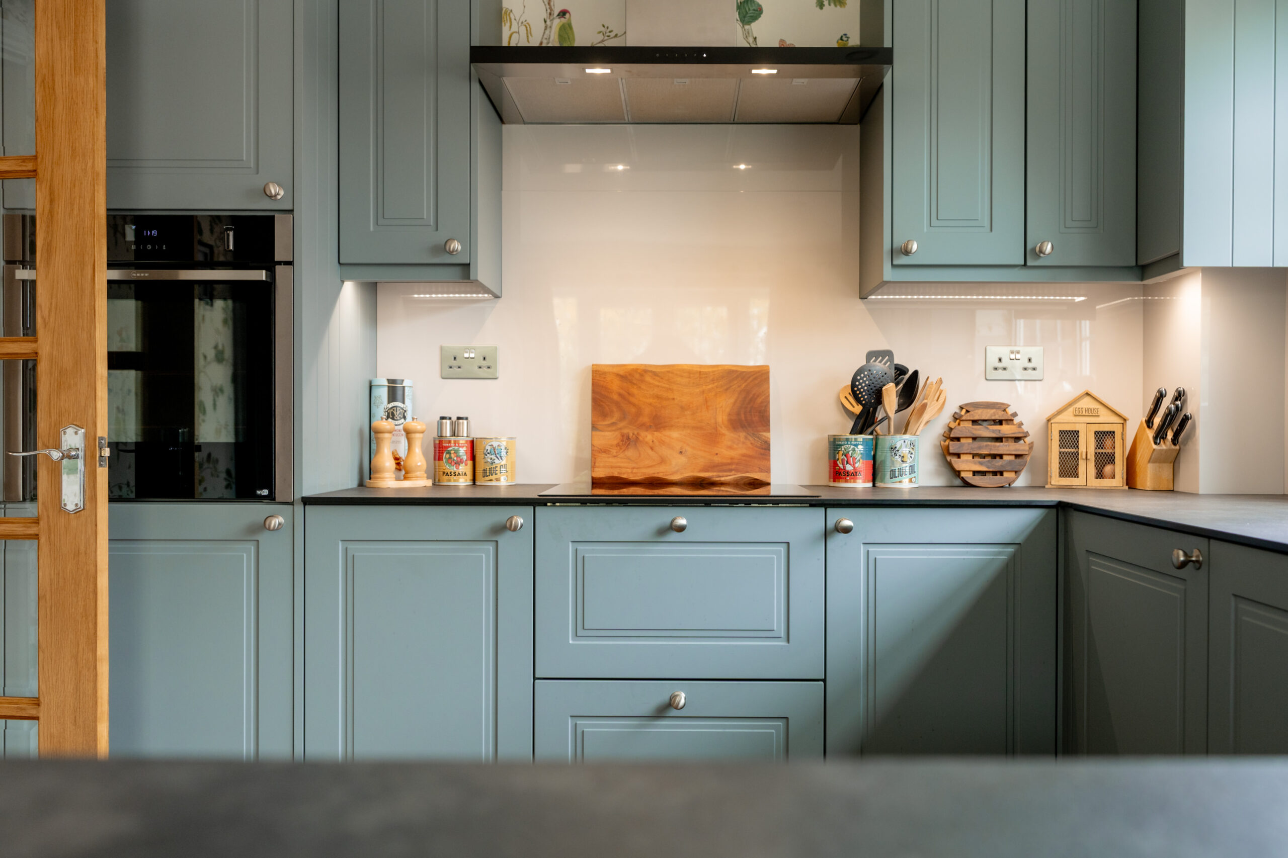 Modern kitchen with light blue cabinets, a wooden cutting board, spice jars, utensils in containers, and small decorative items on the countertop under bright, under-cabinet lighting.