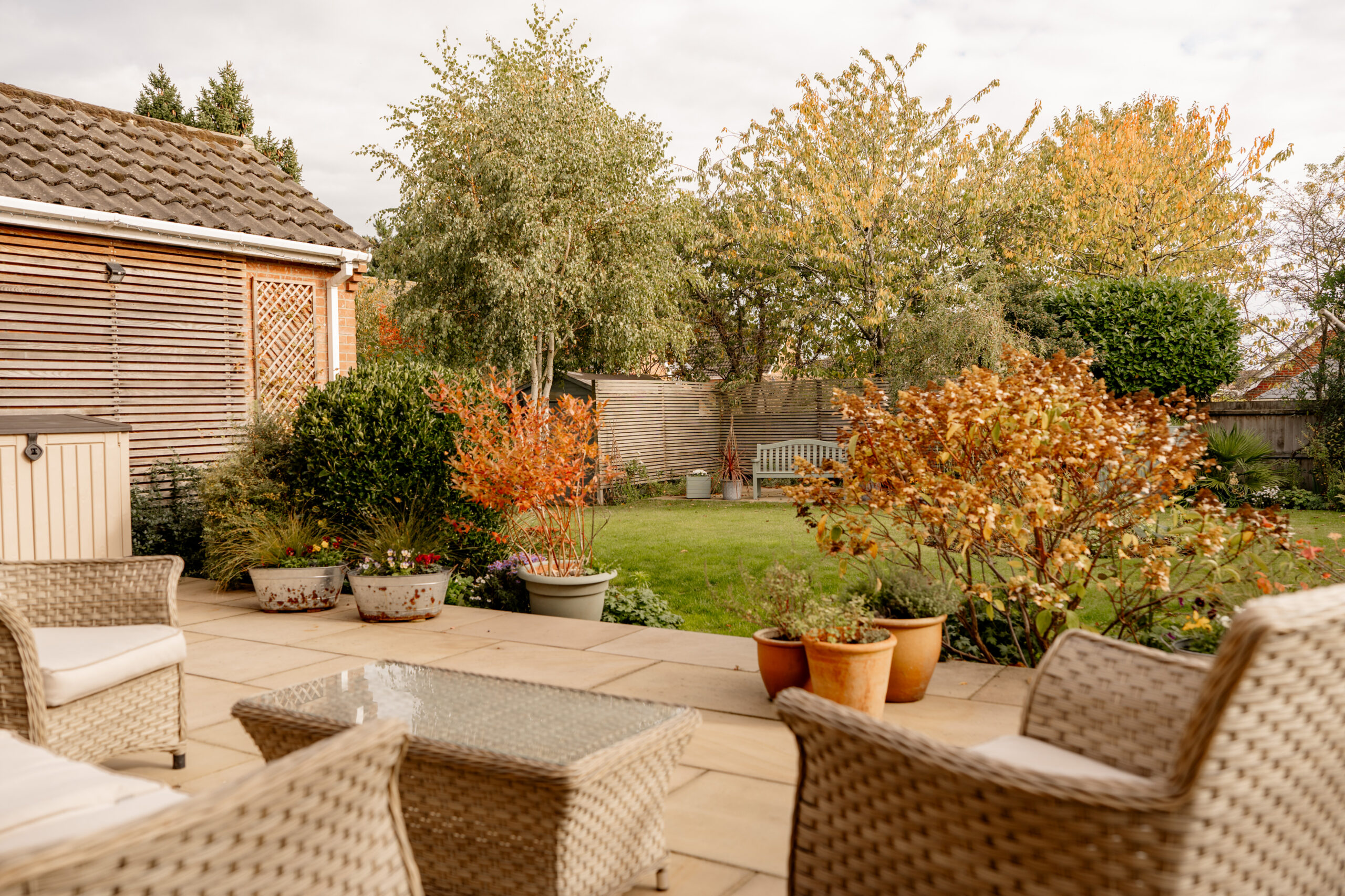 A backyard patio with wicker chairs, a glass-top table, and potted plants overlooks a green lawn bordered by trees and shrubs with autumn foliage. A wooden fence and garden bench are visible in the background.