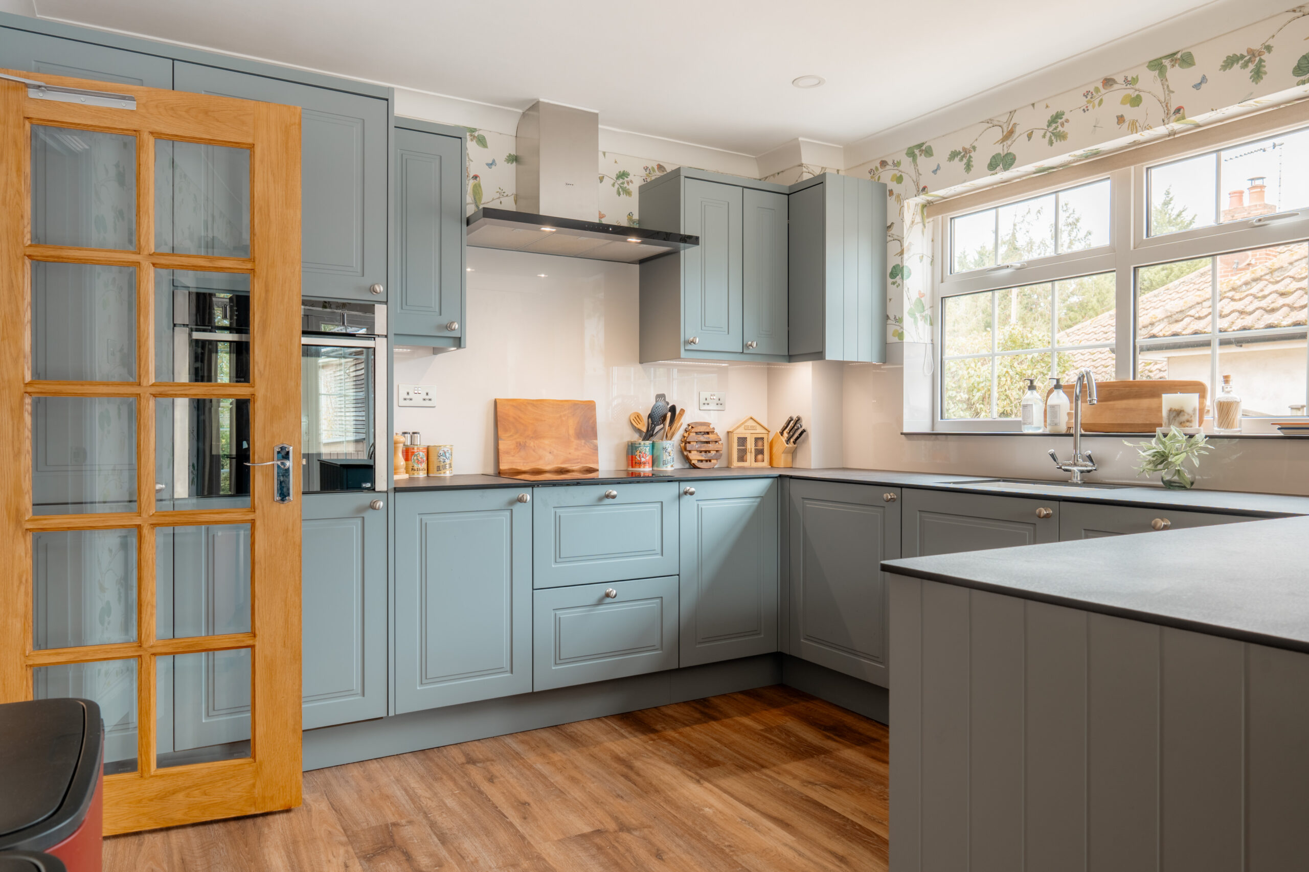 A modern kitchen with light blue cabinets, wooden accents, a built-in oven, and a large window letting in natural light. The floor is wood, and the room appears tidy and inviting.