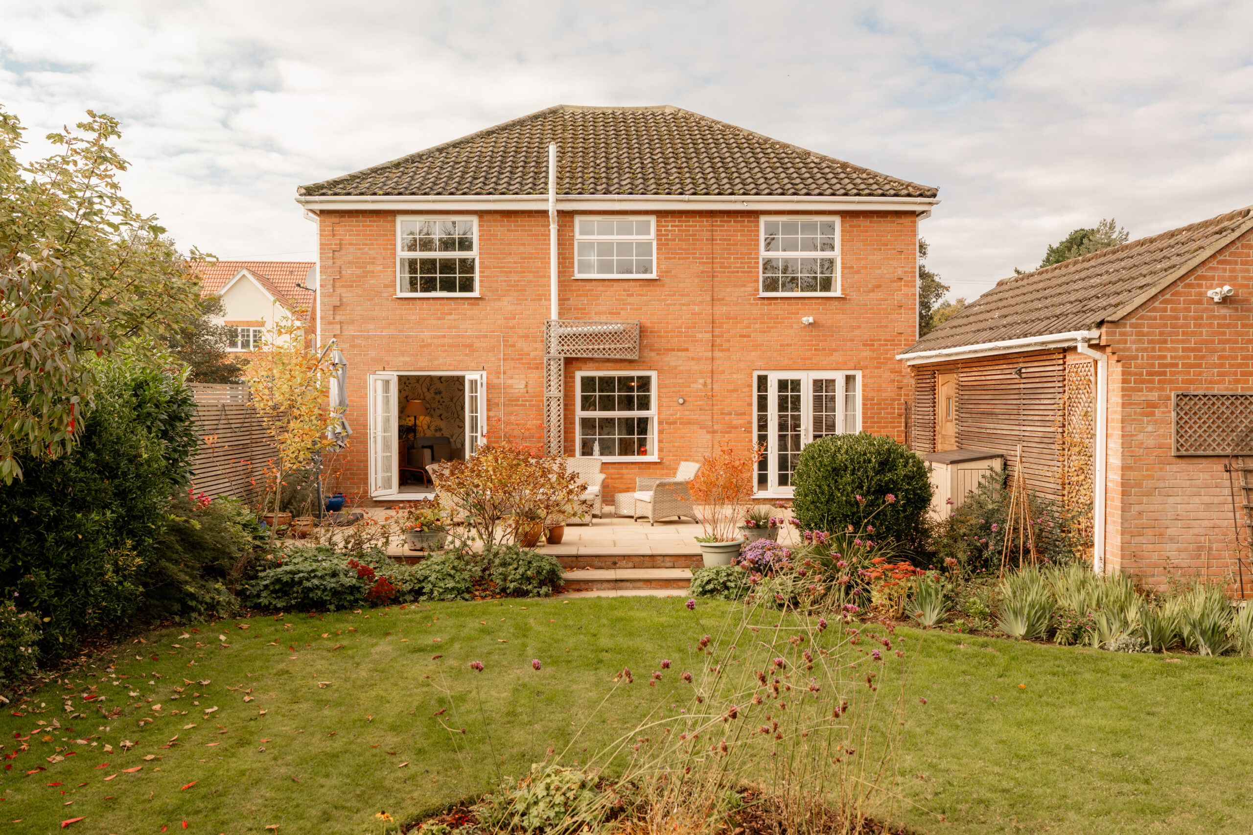 Two-story brick house with large windows and a patio, surrounded by a well-kept garden with shrubs, plants, and a neatly trimmed lawn on a partly cloudy day.