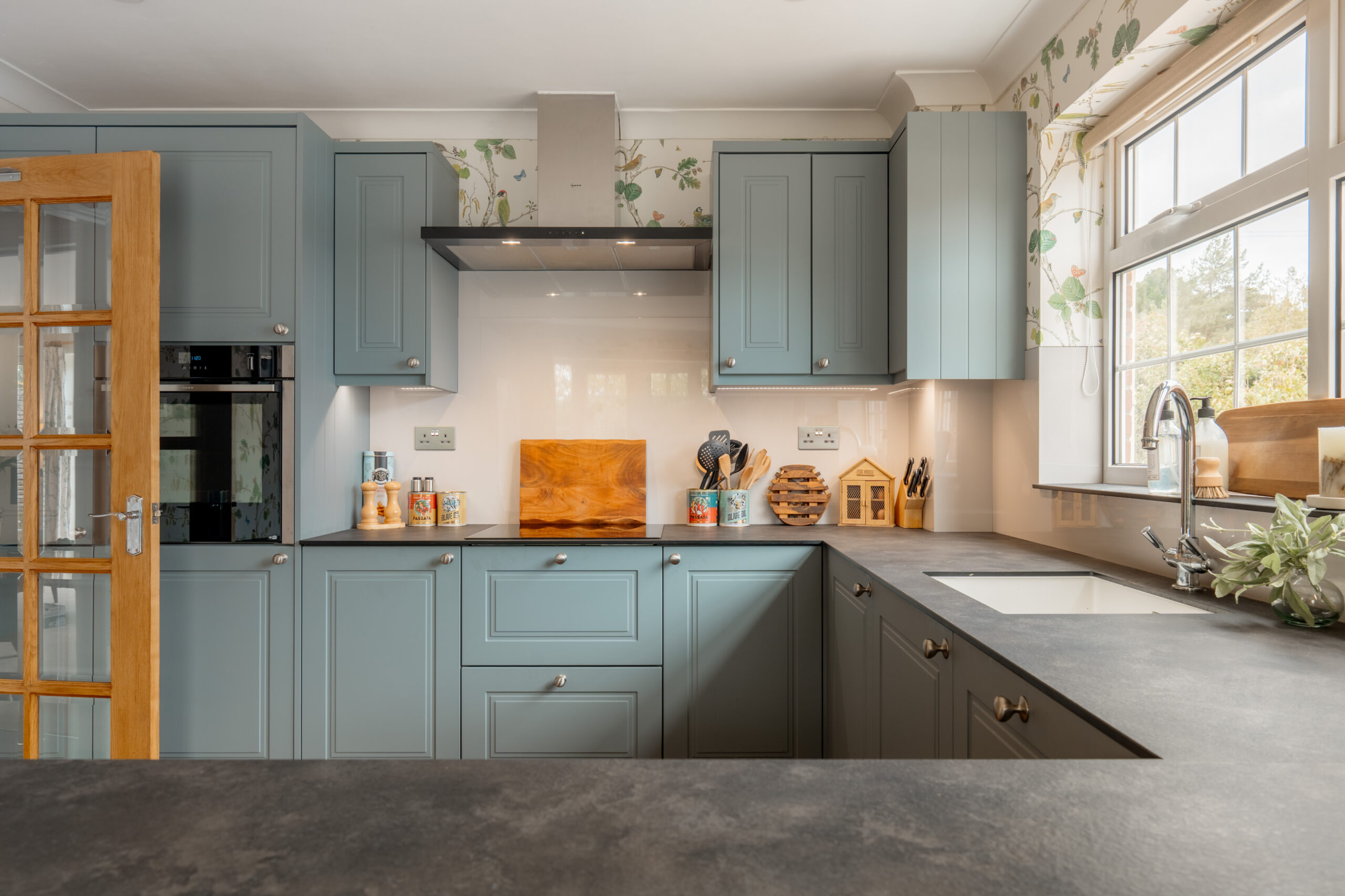 Modern kitchen with light blue cabinets, black countertops, a built-in oven, various utensils, a cutting board, jars, and a window letting in natural light. Light floral wallpaper accents the upper wall.