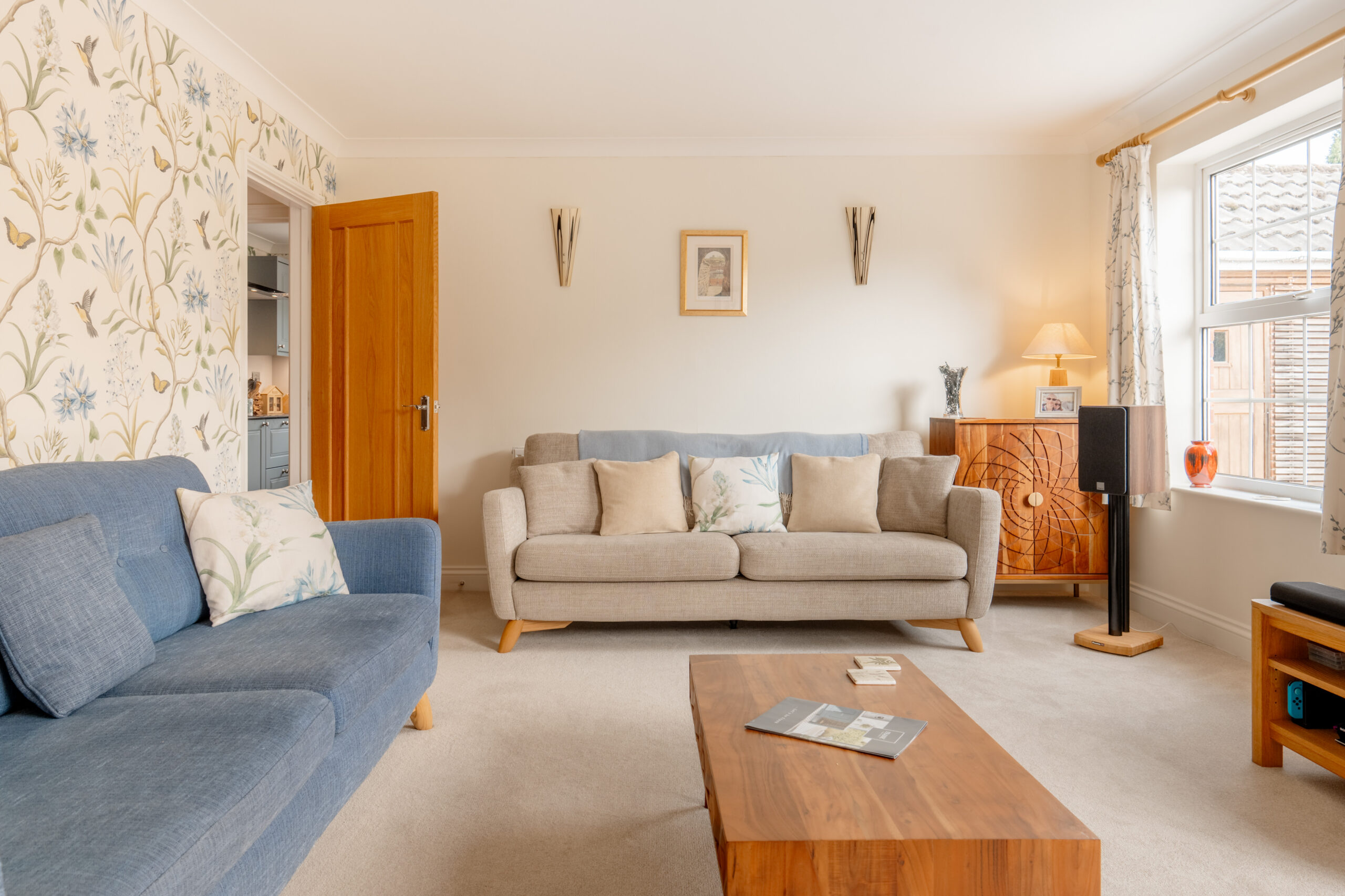 A bright living room with a blue sofa, a beige sofa, a wooden coffee table, and a window letting in natural light. The walls are decorated with floral wallpaper, art, and various decorative items.