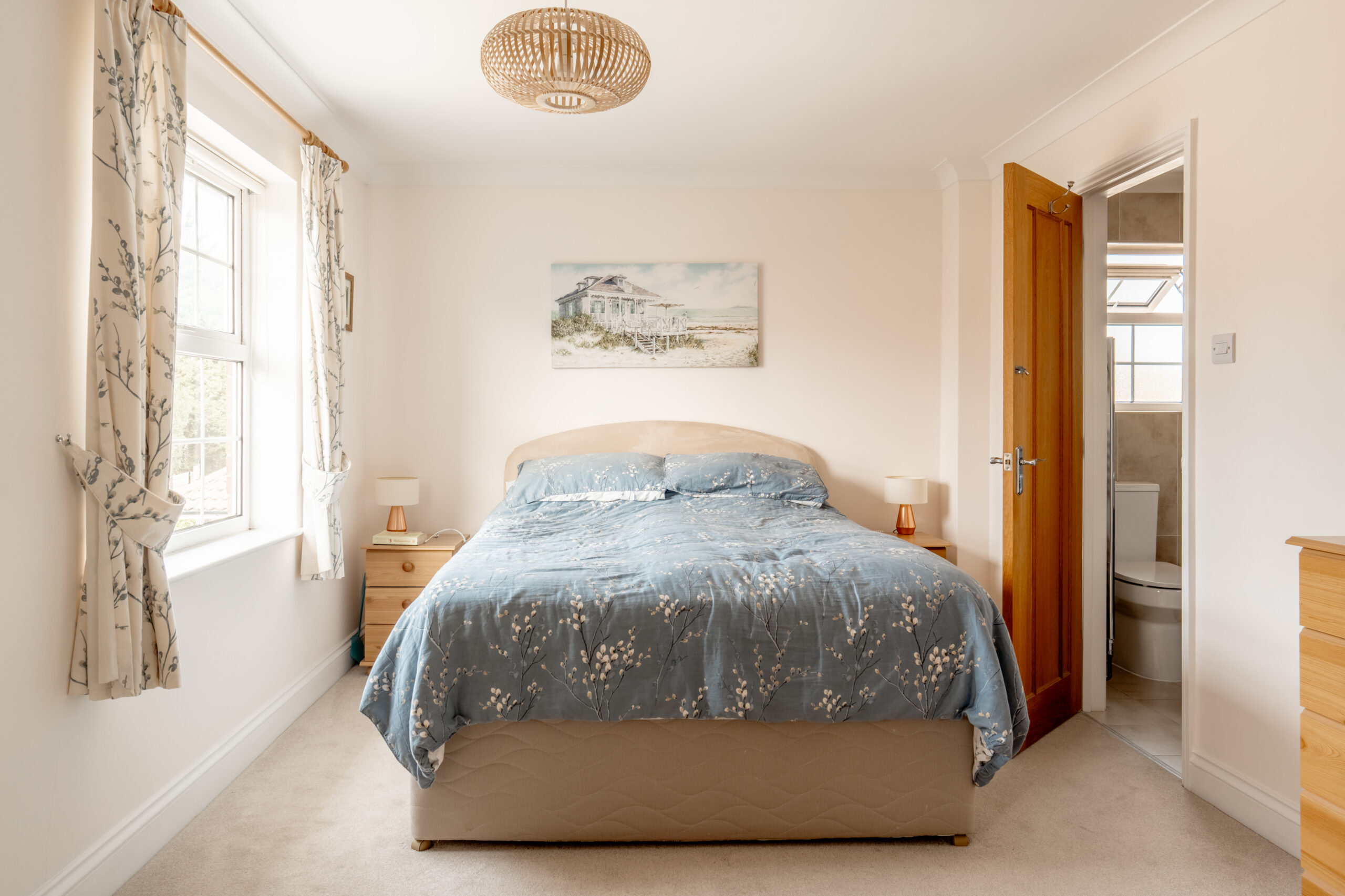 A cozy bedroom with a double bed covered in a blue floral duvet, two wooden nightstands with lamps, a window with floral curtains, and an open door leading to an en-suite bathroom.