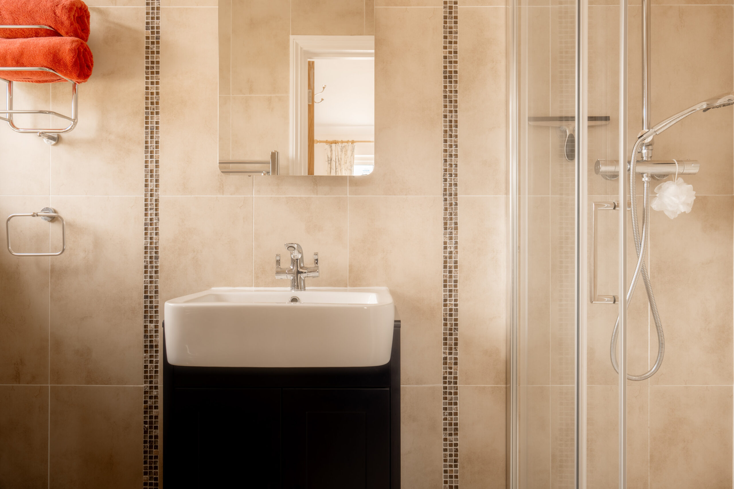 A modern bathroom with beige tiled walls, a white sink with a chrome faucet on a dark cabinet, a wall mirror, a glass shower enclosure, and orange towels on a rack.