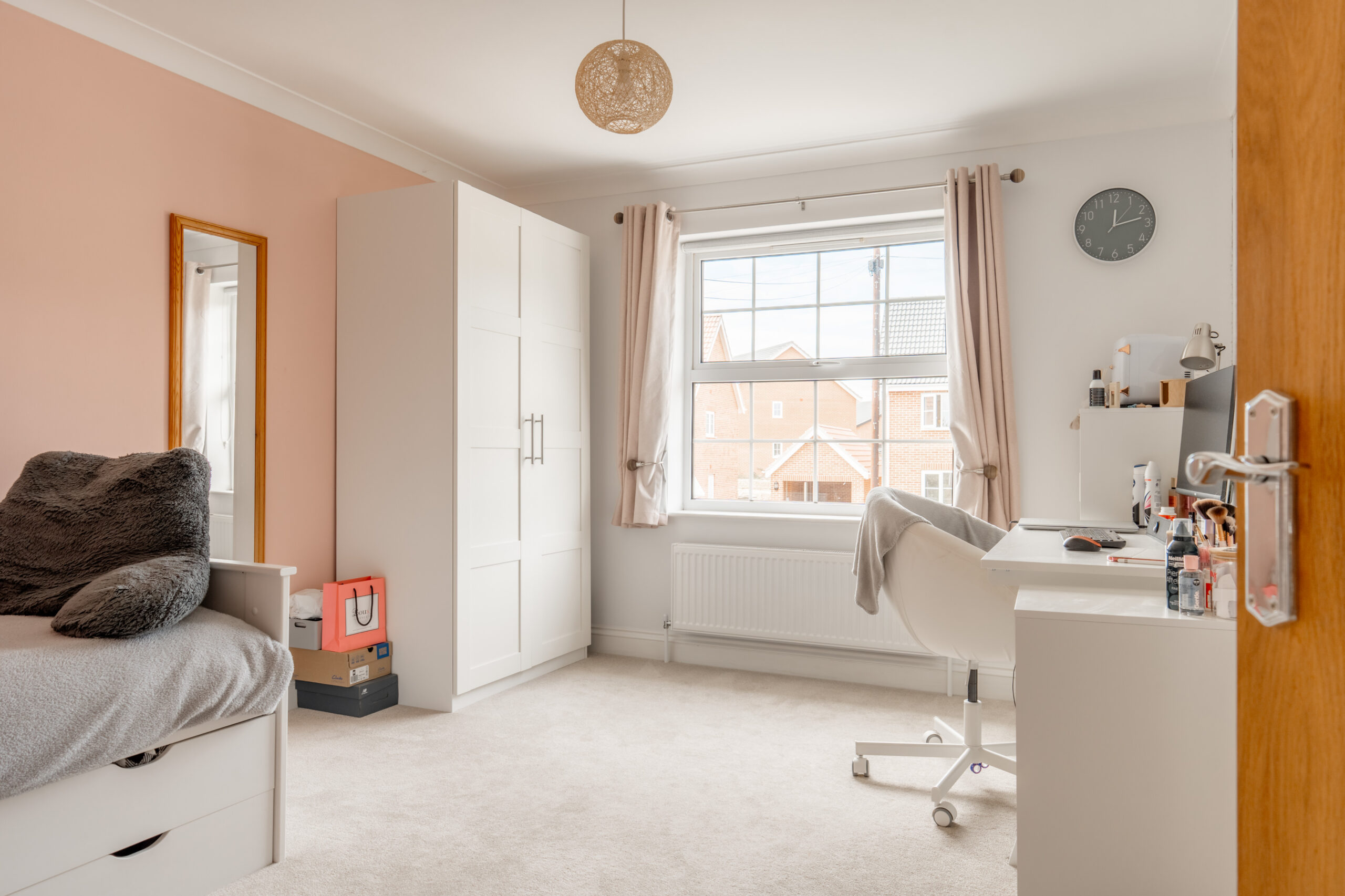 A bright, tidy bedroom with a neatly made bed, a white wardrobe, a desk with a white chair, a wall clock, and a large window with light curtains letting in natural light.