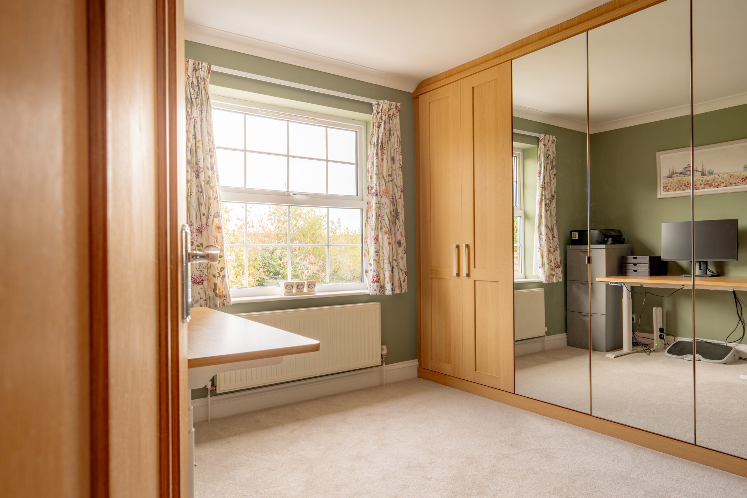 Bright bedroom with green walls, large mirrored wardrobe, beige carpet, floral curtains, and a window letting in natural light. A desk with a computer is on the right side near the window.