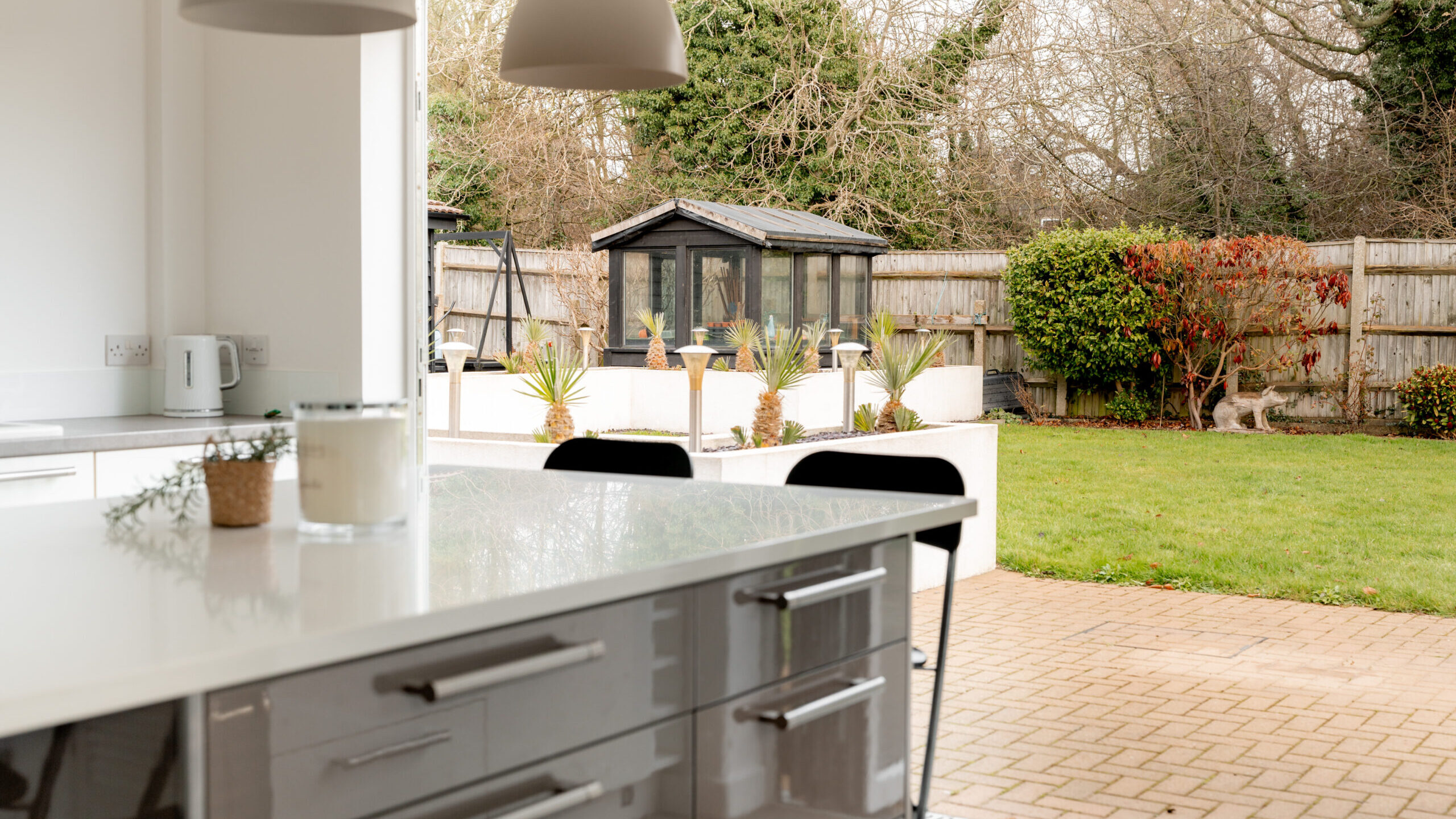 Modern kitchen with a glossy countertop and pendant lights, opening onto a well-kept backyard with green grass, a paved area, plants, and a dark garden shed along a wooden fence.