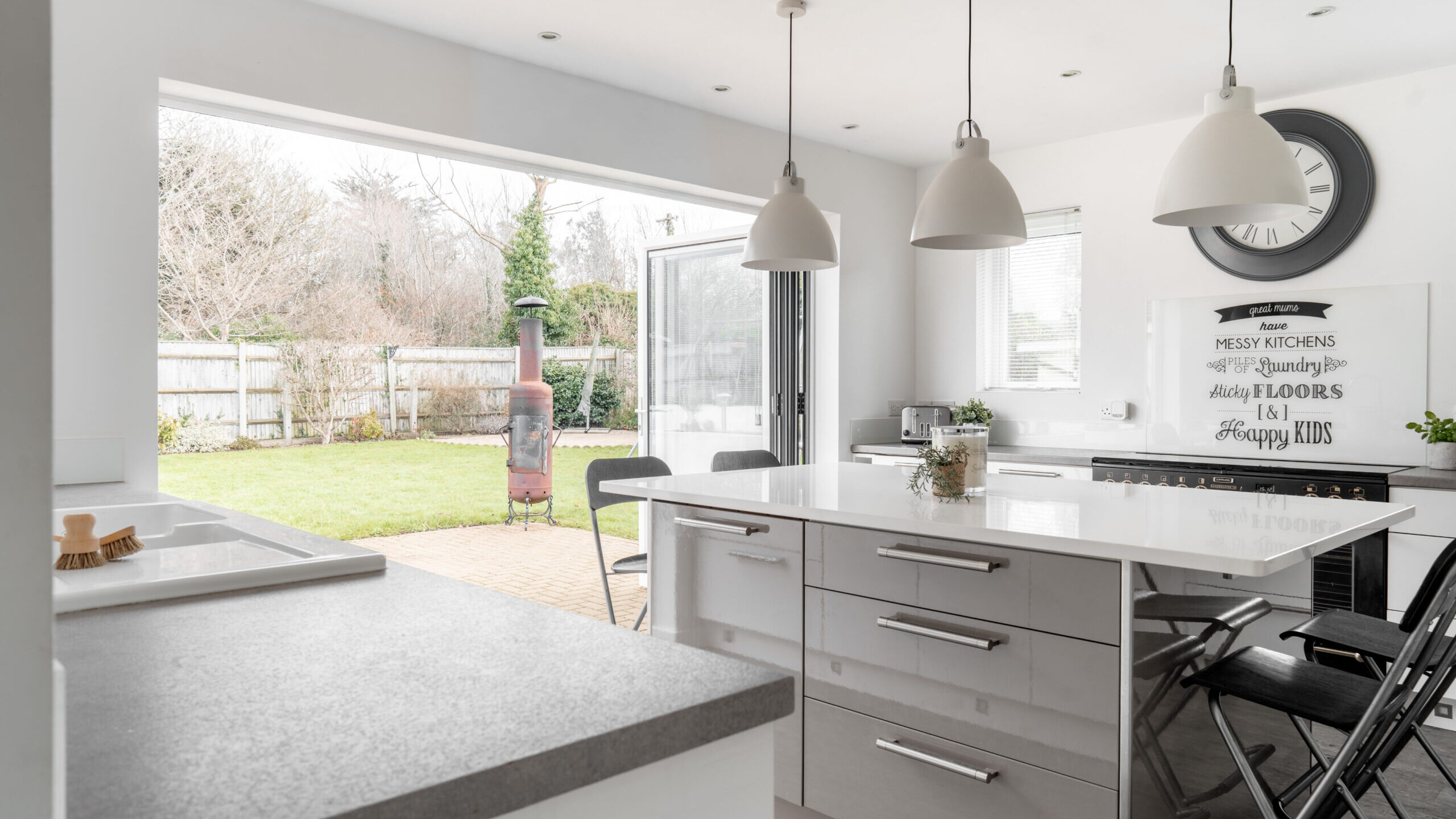 A bright modern kitchen with white cabinets and countertops, three pendant lights, and bar stools. Glass doors open to a patio and green garden, with an outdoor wood-burning stove visible.