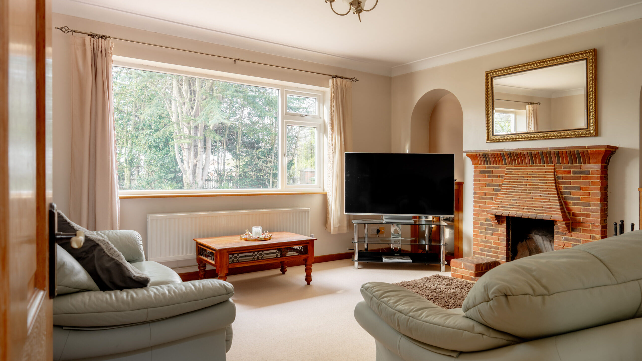 A cozy living room with cream walls, two light leather sofas, a wooden coffee table, a large window with beige curtains, a brick fireplace with a mirror above it, and a flat-screen TV on a stand.