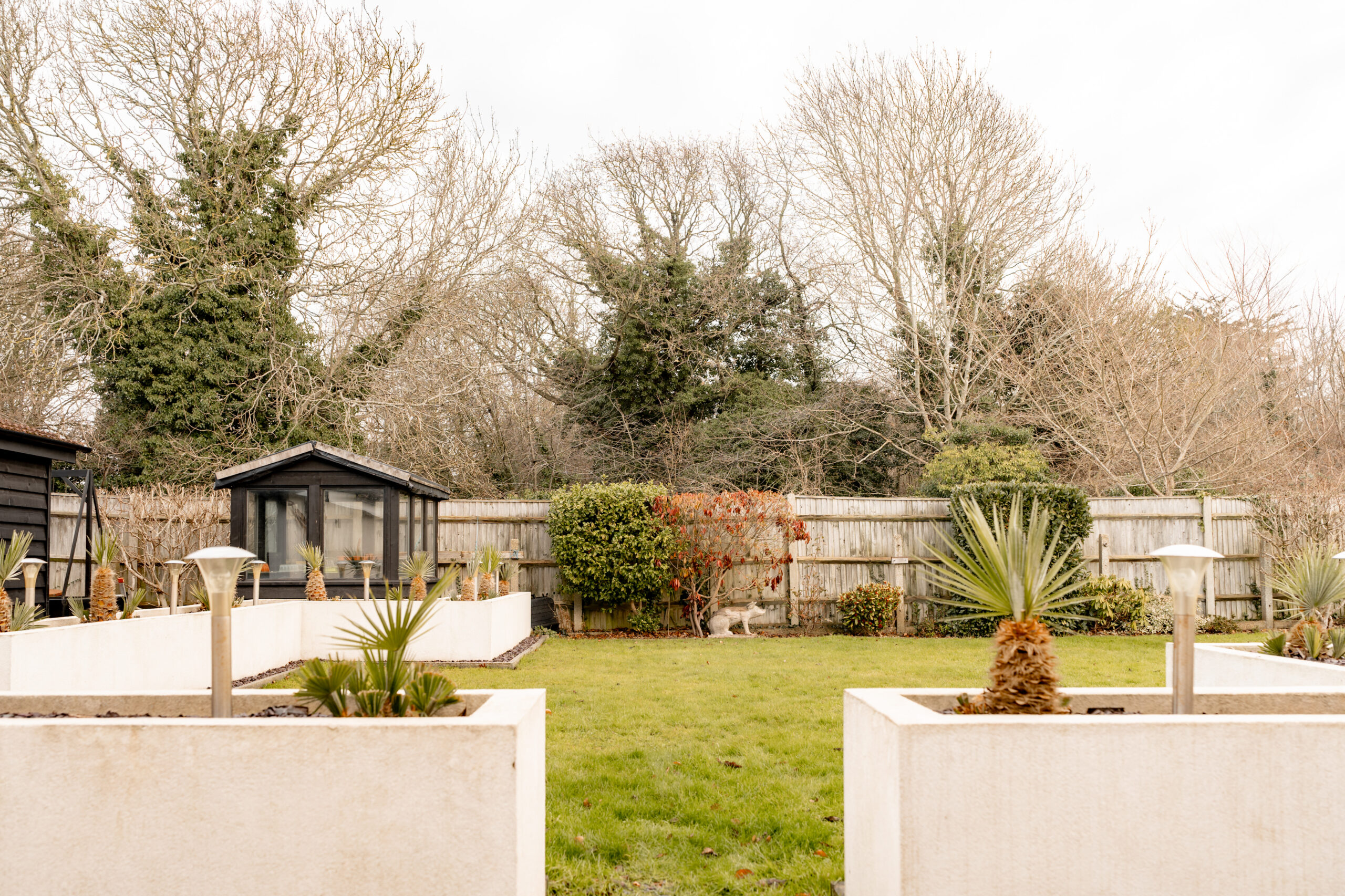 A neatly landscaped garden with a manicured lawn, potted plants in white planters, a small garden shed, and a wooden fence. Leafless trees and dense greenery are visible beyond the fence under an overcast sky.