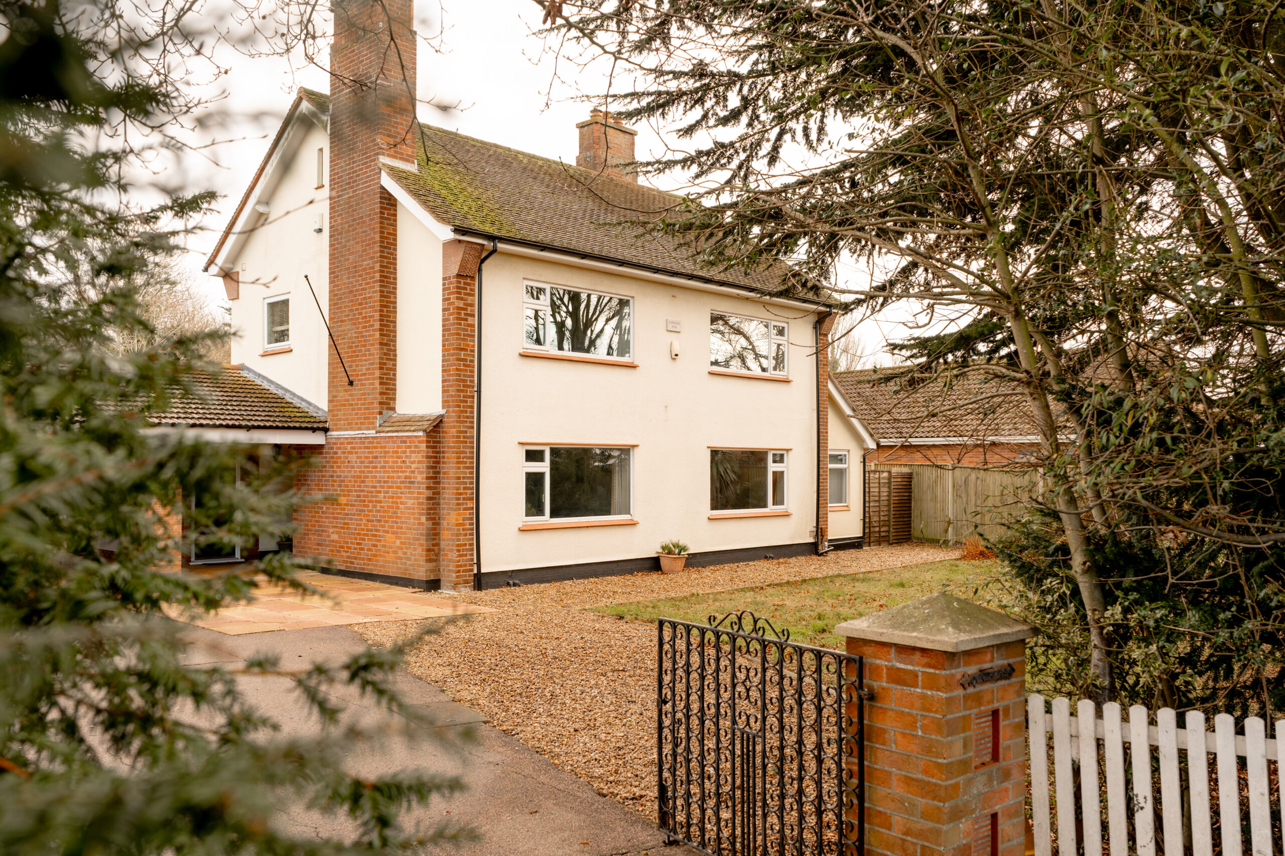 A two-story detached house with white walls, red brick accents, and a tiled roof, set behind a black metal gate and white picket fence, surrounded by trees and a gravel driveway.