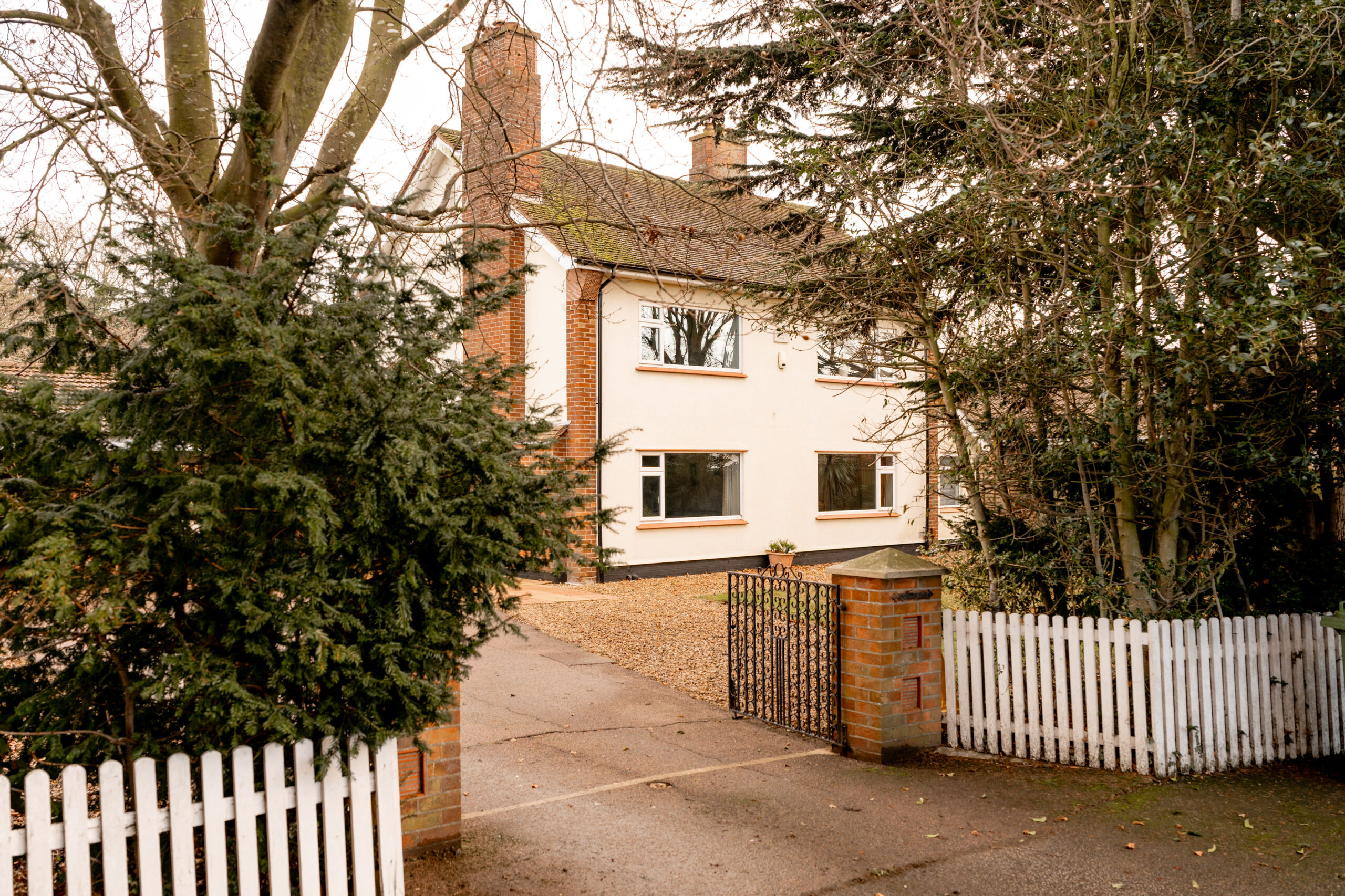 A two-story white house with a sloped roof sits behind a white picket fence and metal gate, surrounded by trees and bushes. The driveway is lined with gravel, leading up to the entrance.