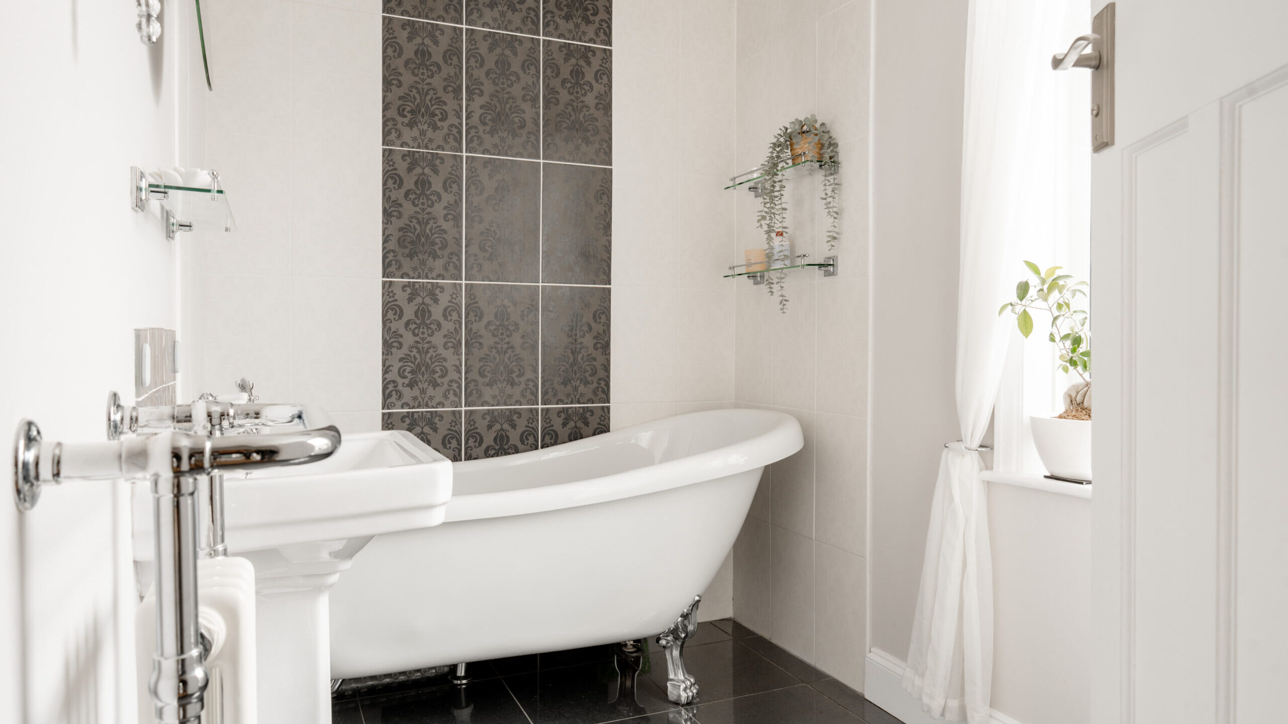 A modern bathroom with a white clawfoot bathtub, a patterned tile accent wall, floating glass shelves with plants, a white sink, and a window with sheer curtains letting in natural light.