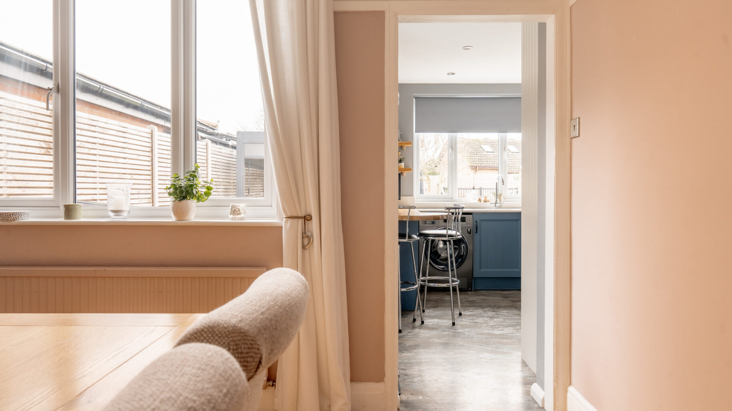 Bright dining room with beige walls and a wooden table, looking through an open doorway into a modern kitchen with blue cabinets and a breakfast bar. Large windows let in natural light.