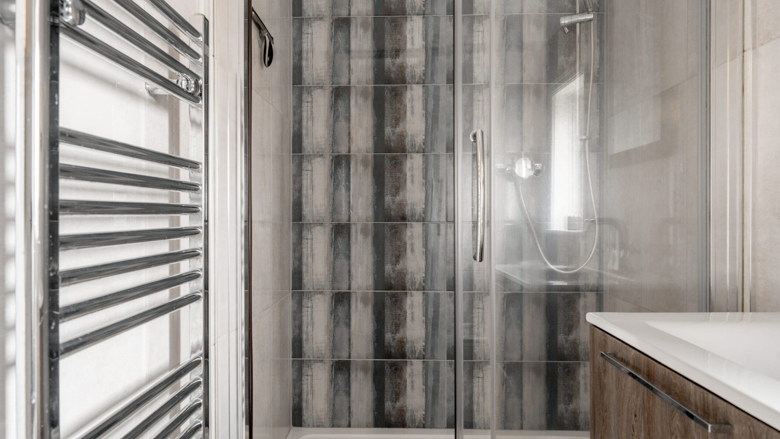 Modern bathroom with a glass shower enclosure, patterned gray and beige tiles on the shower wall, a chrome towel warmer, and a wooden vanity with a white sink.
