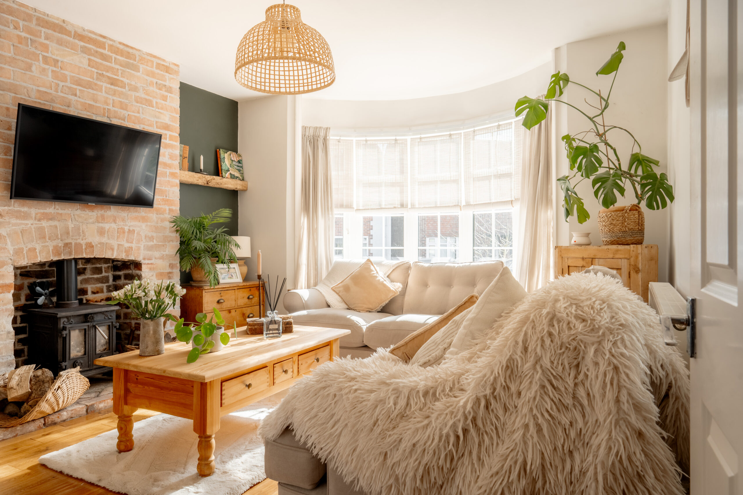 Bright, cozy living room with a curved bay window, beige sofa, wooden coffee table, indoor plants, TV above a brick fireplace, and a fluffy throw over a chair; natural light fills the space.