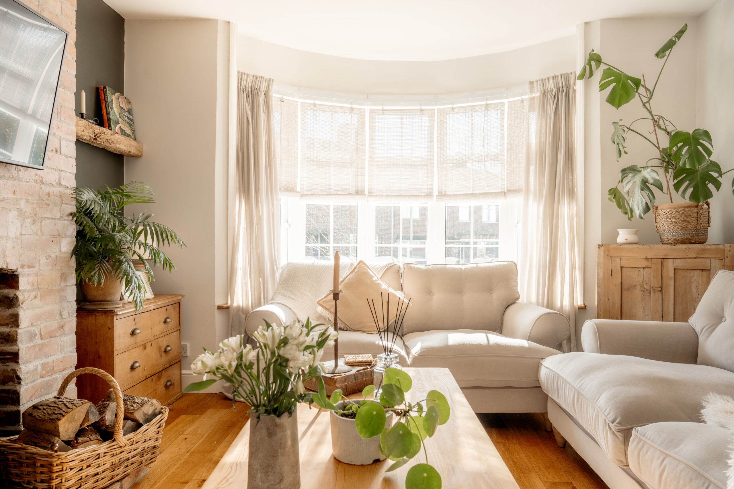 A bright, cozy living room with cream sofas, a wooden coffee table with plants, a bay window with sheer curtains, wooden furniture, a potted monstera, and sunlight streaming in.