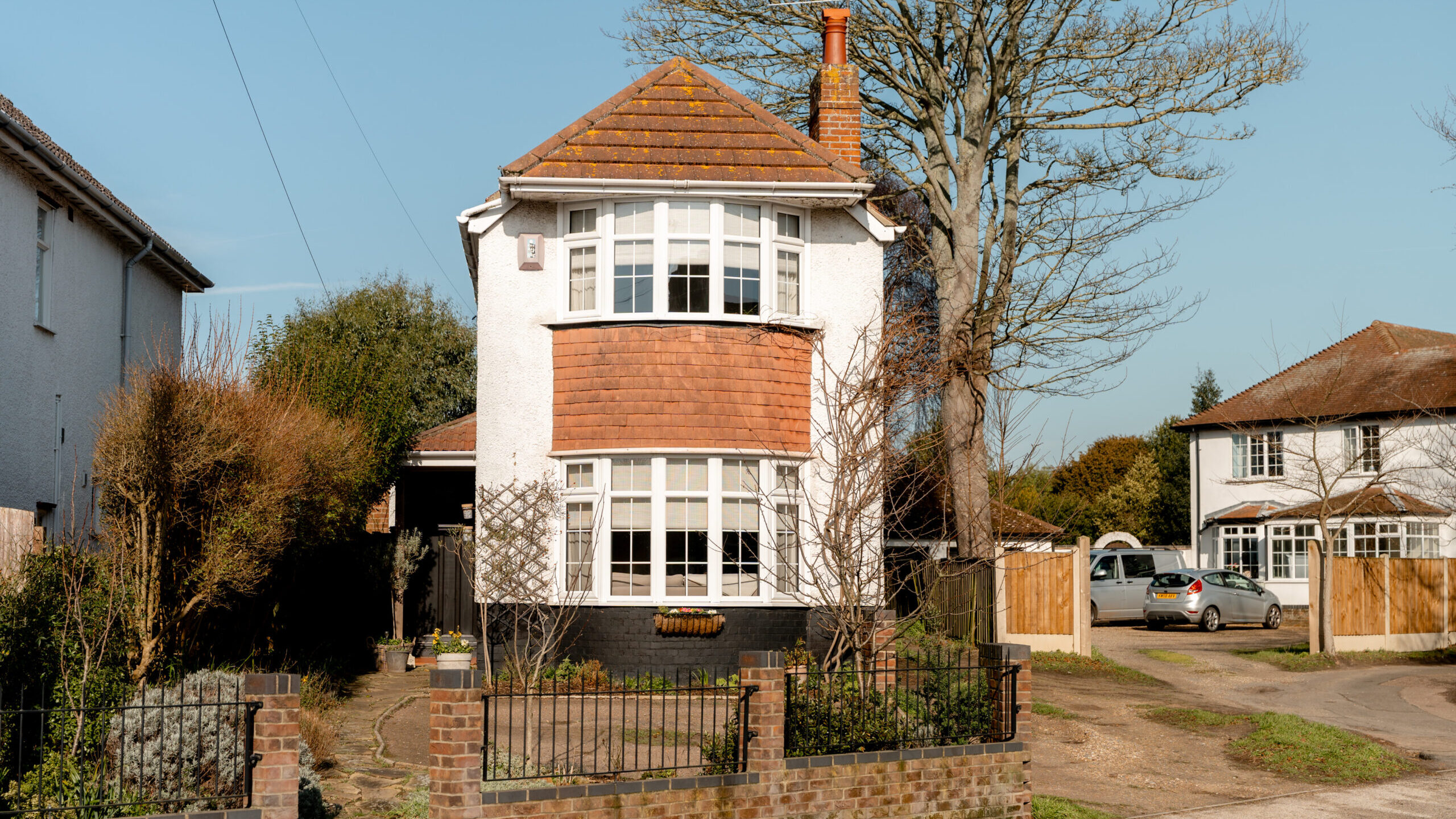 A two-story detached house with white walls, a red-tiled roof, and large bay windows, surrounded by a low brick wall and a front garden. A tree stands beside the house on a sunny day.