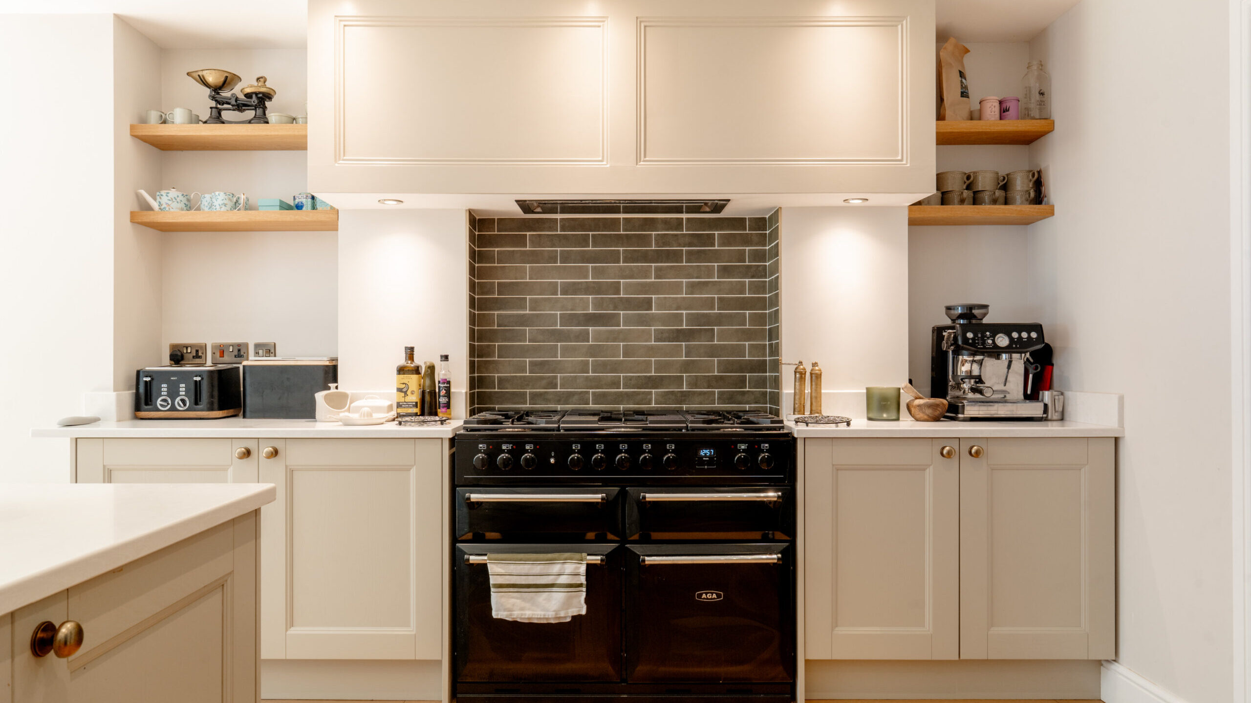 A modern kitchen with cream cabinets, a black double oven, a gray subway tile backsplash, wooden shelves holding dishes and glasses, a toaster, scale, and a coffee machine on the white countertops.