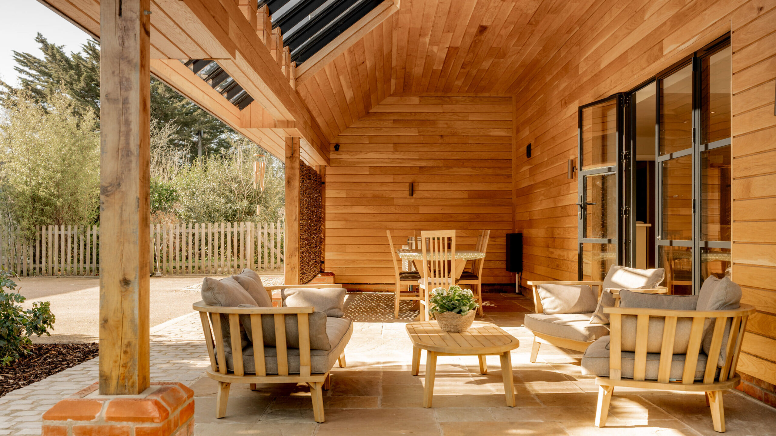 A cozy covered patio with wooden walls and ceiling, cushioned wooden chairs, a round coffee table with a plant, and a dining table in the background, all bathed in warm natural light.