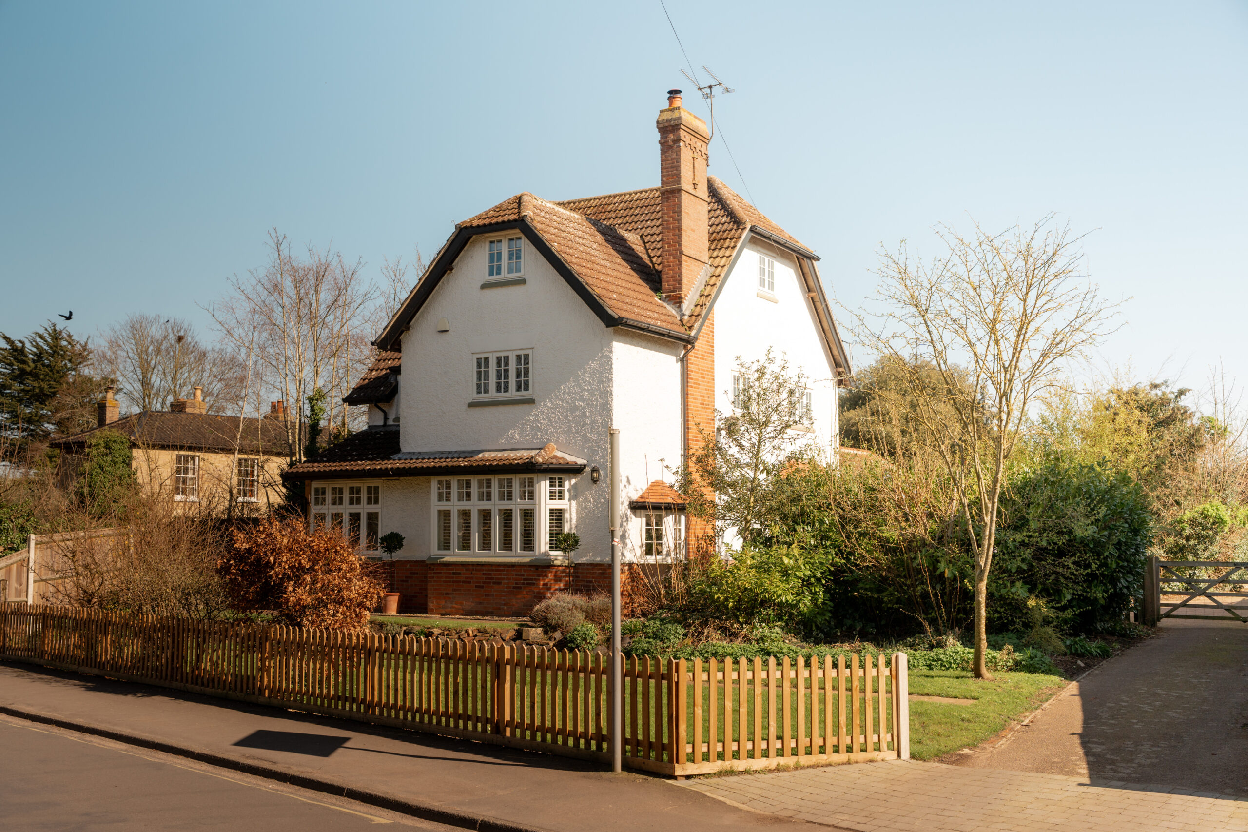 A two-story white house with a red-tiled roof, surrounded by greenery and a wooden picket fence, sits on a sunny day next to a quiet street.