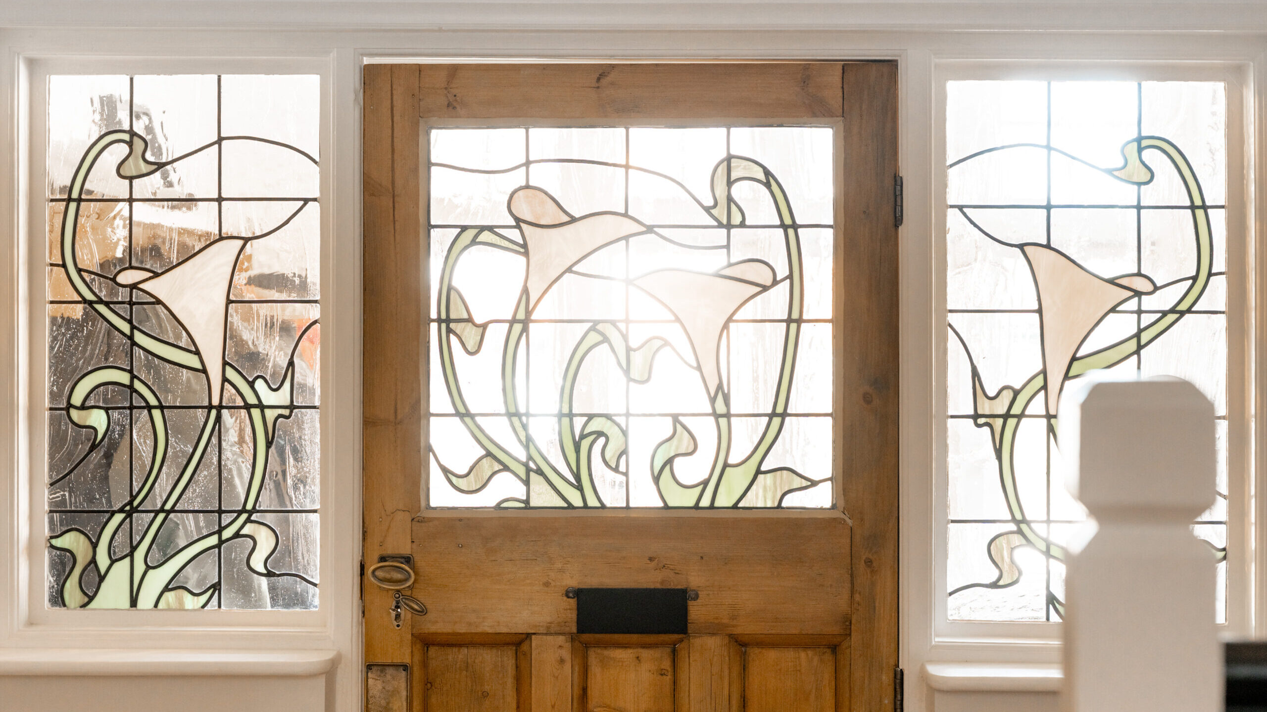 A wooden door with two side windows, all featuring stained glass designs of white calla lilies and green leaves. Sunlight shines through, illuminating the floral patterns.