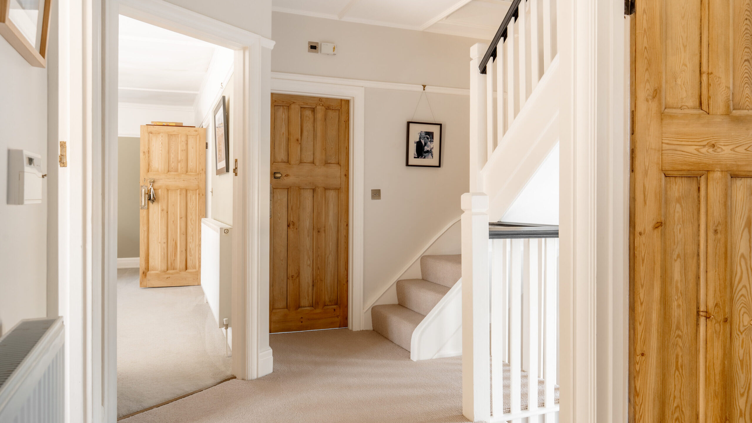 Bright hallway in a home with beige carpet, wooden doors, white walls, and a staircase with white railings. One room visible on the left with an open door and a wardrobe inside. A framed picture hangs near the stairs.