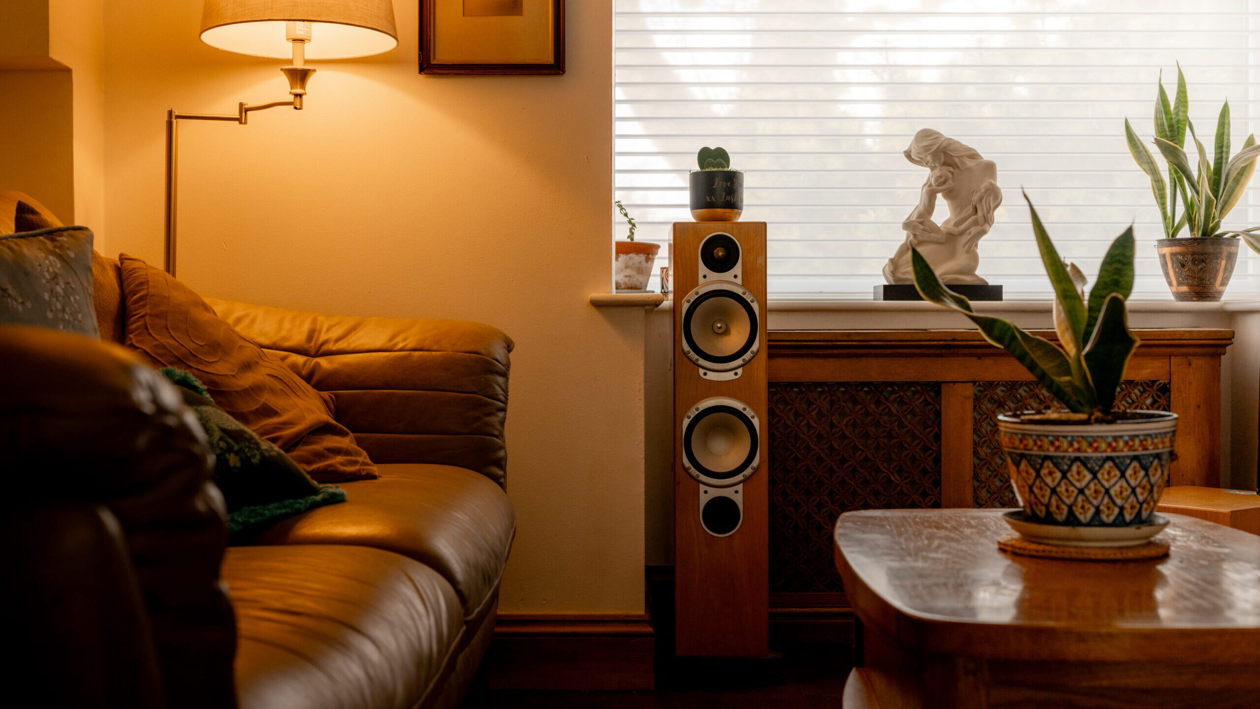 A cozy living room with a brown leather sofa, a wooden coffee table with a potted plant, a floor lamp, a speaker, and a windowsill holding another plant and a sculpture. Warm lighting creates a relaxed atmosphere.
